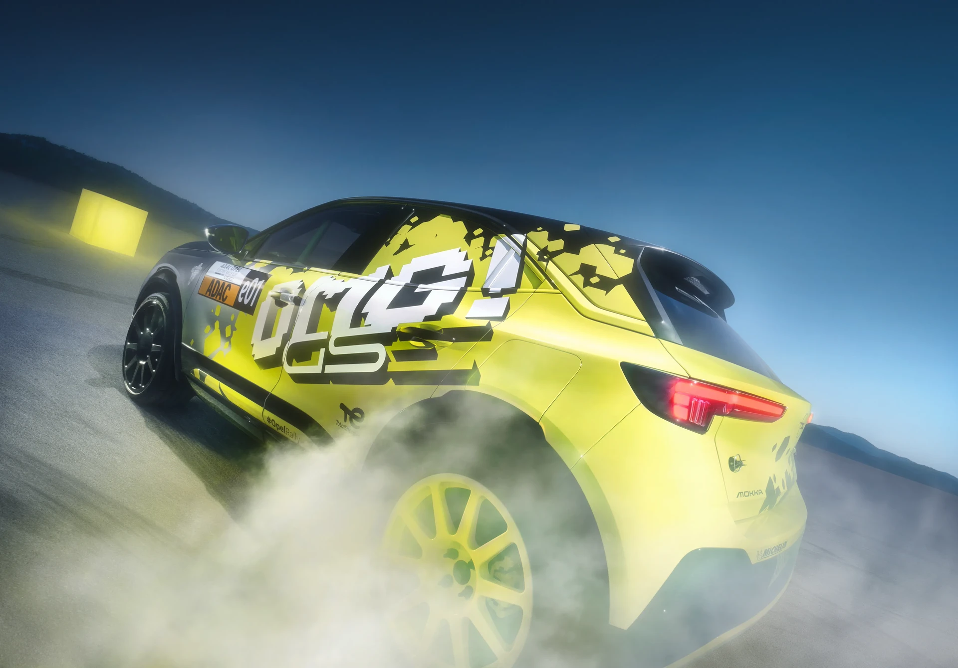 Opel Mokka GSe Rallyeauto in gelber Rennlackierung beim Driften auf einer Strecke mit Reifenrauch, Heck-Dreiviertelansicht vor blauem Himmel.