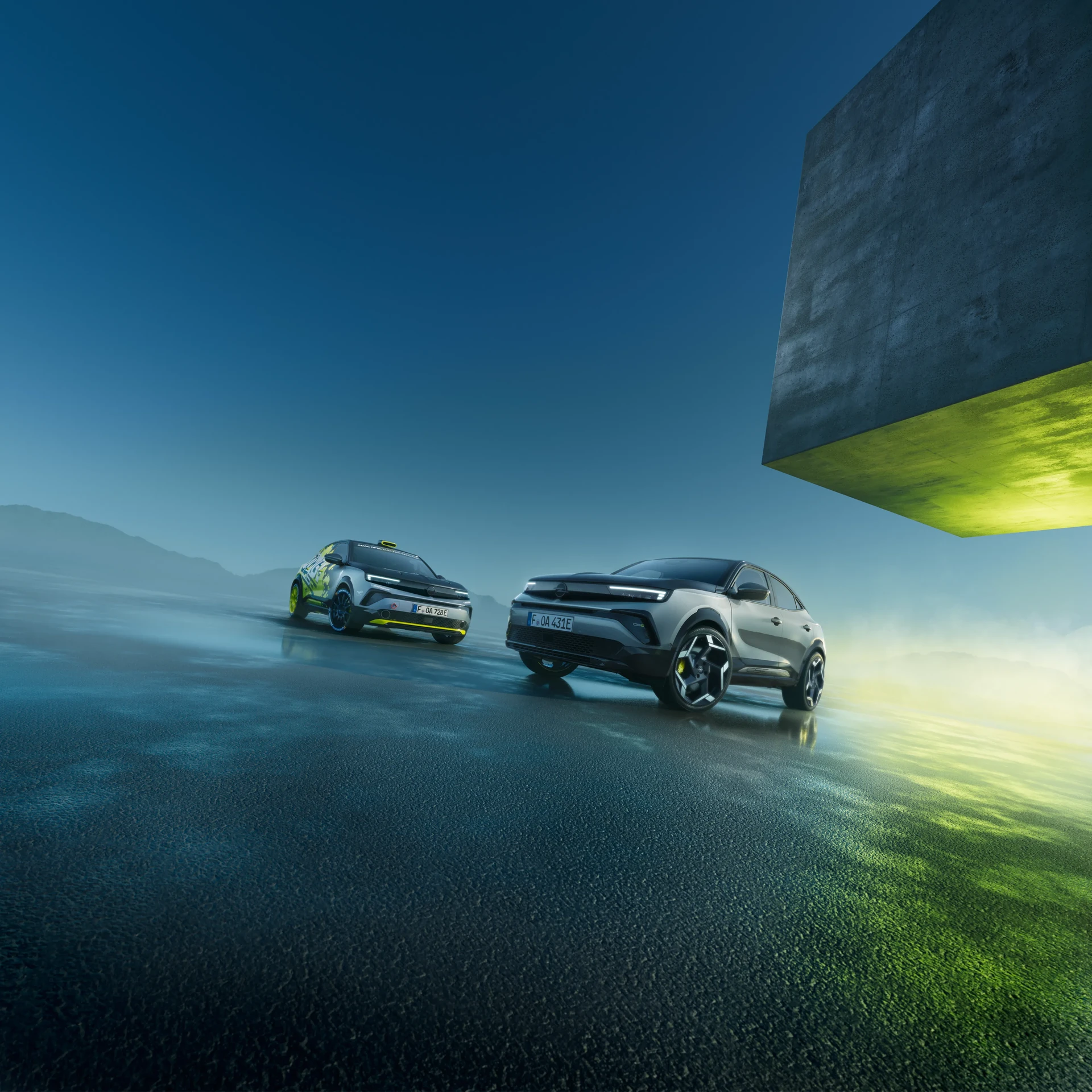 Zwei Opel Mokka GSe Elektrofahrzeuge nebeneinander auf einer reflektierenden Oberfläche — eines in Rallye-Rennlackierung und eines in serienmäßigem Grau — unter einem dramatischen blauen Himmel mit einem Betonarchitekturelement darüber.