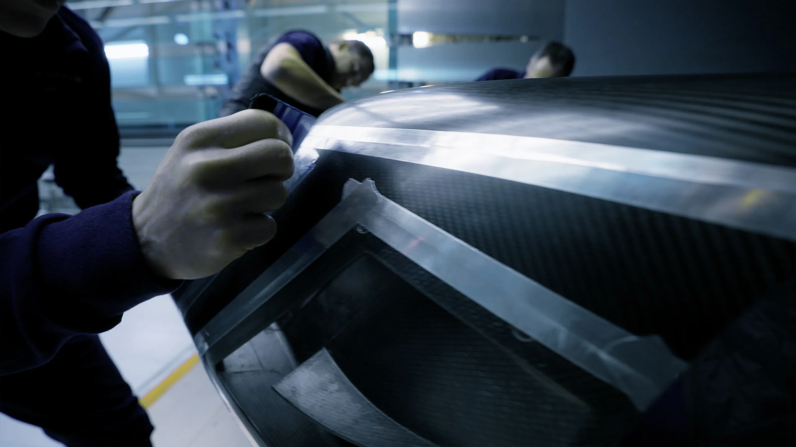 Close-up of hands applying masking tape along the edge of a carbon fibre body panel on a race car, with two team members working in the blurred background of the workshop.