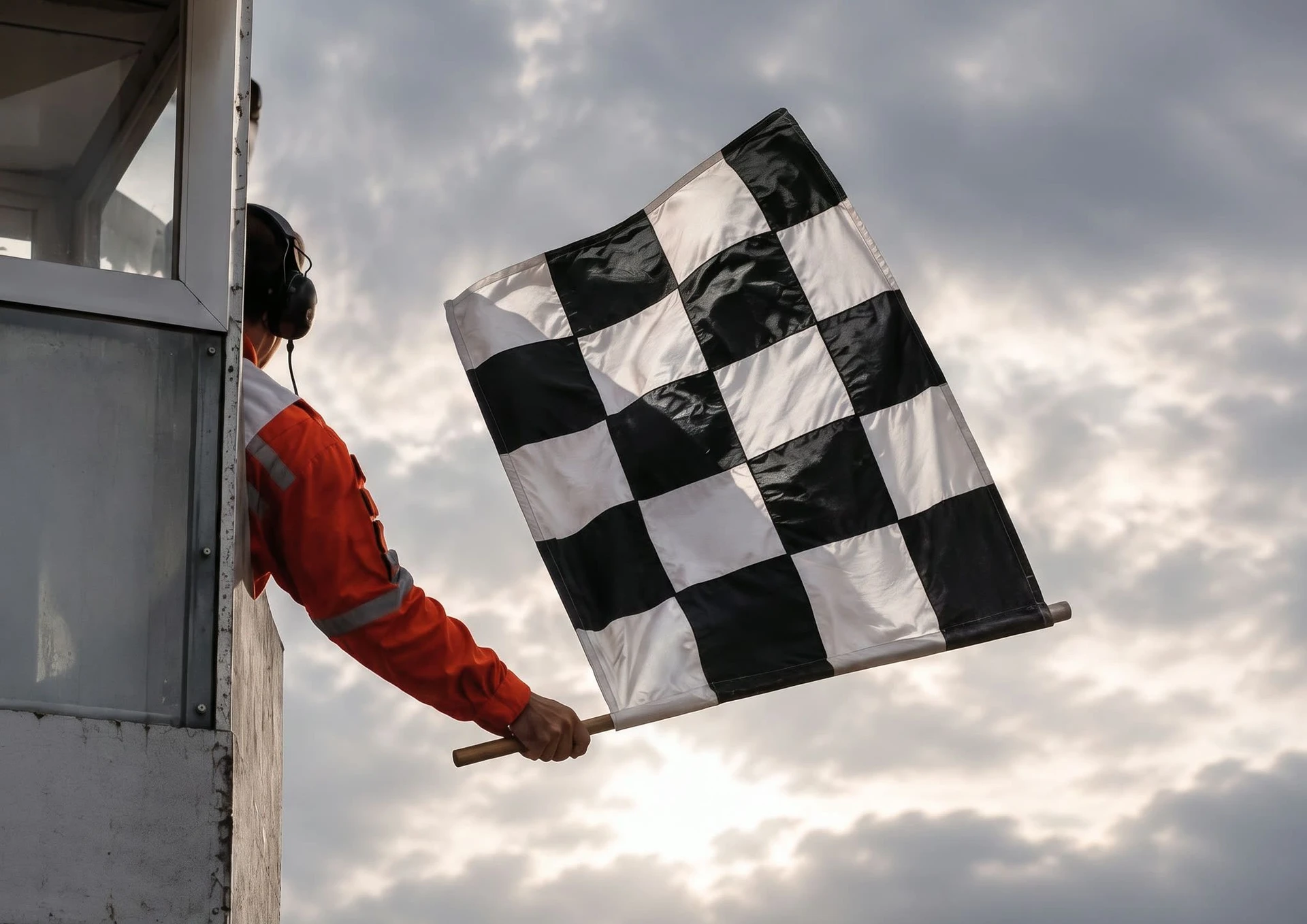 A race marshal in an orange suit and ear defenders leaning out from the start-finish gantry to wave the chequered flag, with empty grandstands blurred below.