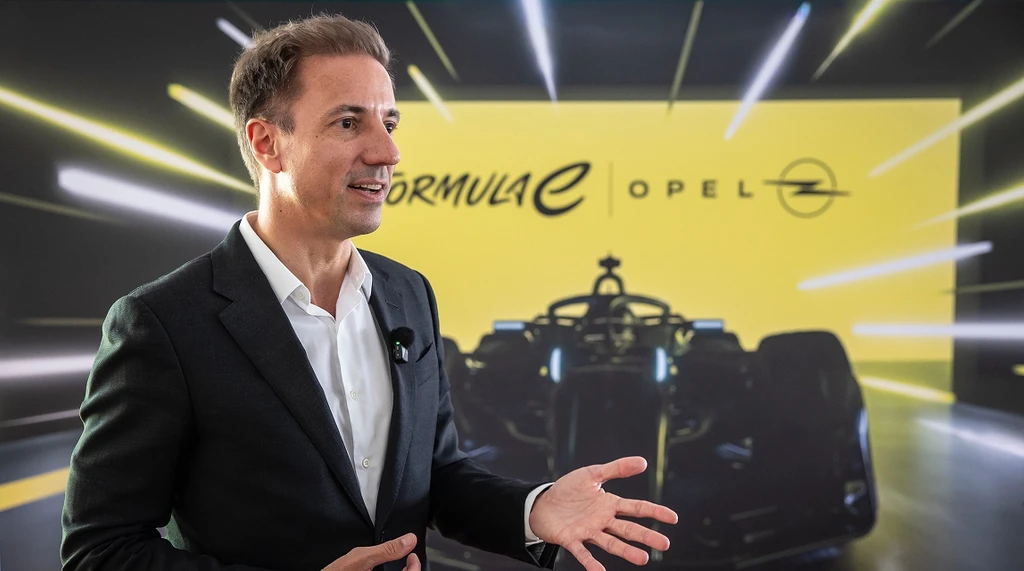 Florian Huettl, CEO der Opel Automobile GmbH, in dunklem Anzug und weißem Hemd, spricht bei einer Opel-Formel-E-Veranstaltung und gestikuliert mit seiner rechten Hand. Im Hintergrund zeigt ein gelbes Werbebanner die Logos von Formel E und Opel sowie ein dunkles Formel-E-Rennfahrzeug.