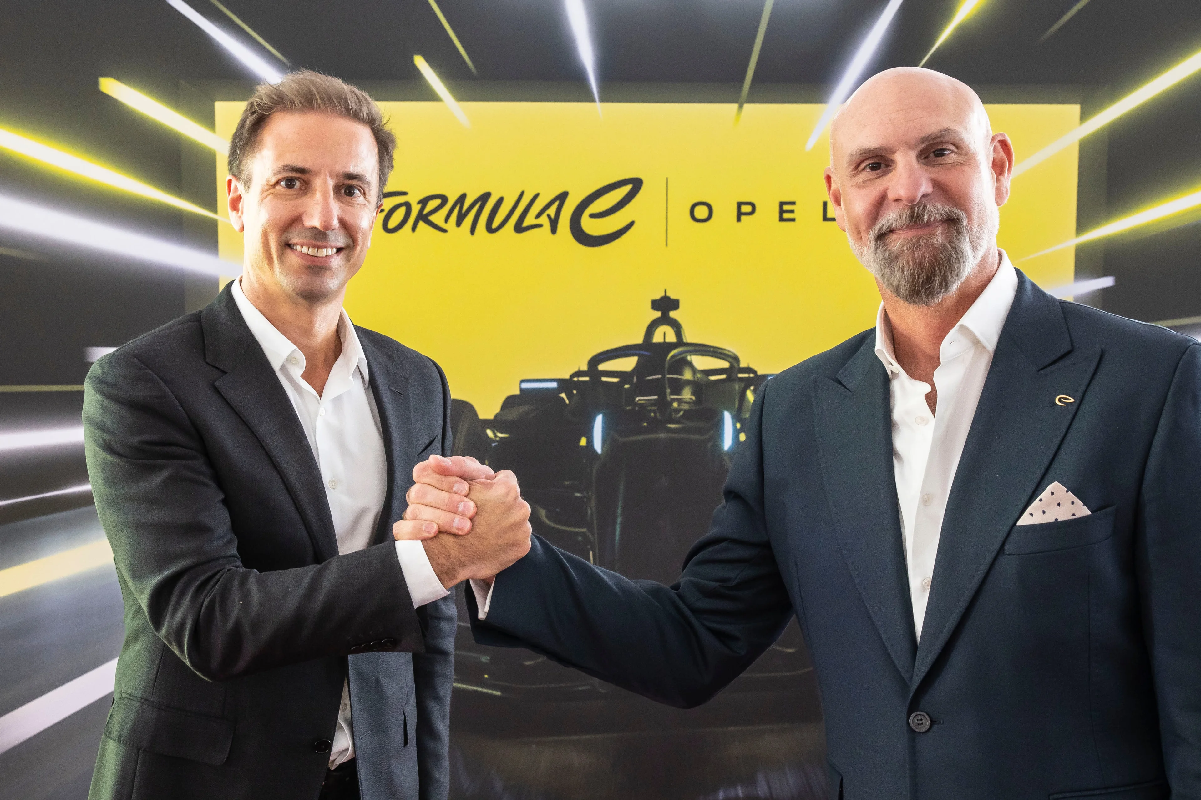 Two men in dark suits shaking hands and smiling at the camera in front of a yellow backdrop showing the Formula E and Opel logos side by side, with a single-seater race car silhouette below.
