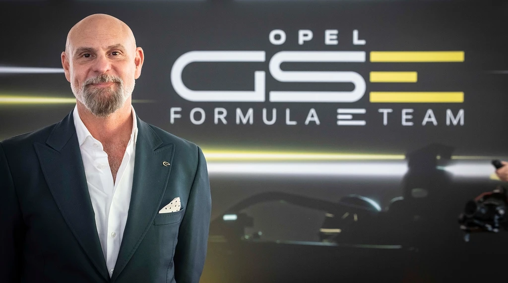 Jeff Dodds, CEO der Formel E, gekleidet in einen dunklen Anzug und weißes Hemd mit Einstecktuch, steht vor einem dunklen Hintergrund mit dem Logo des ‚Opel GSe Formula E Team' in weißer und gelber Schrift. Im Hintergrund ist ein Formel-E-Fahrzeug teilweise sichtbar