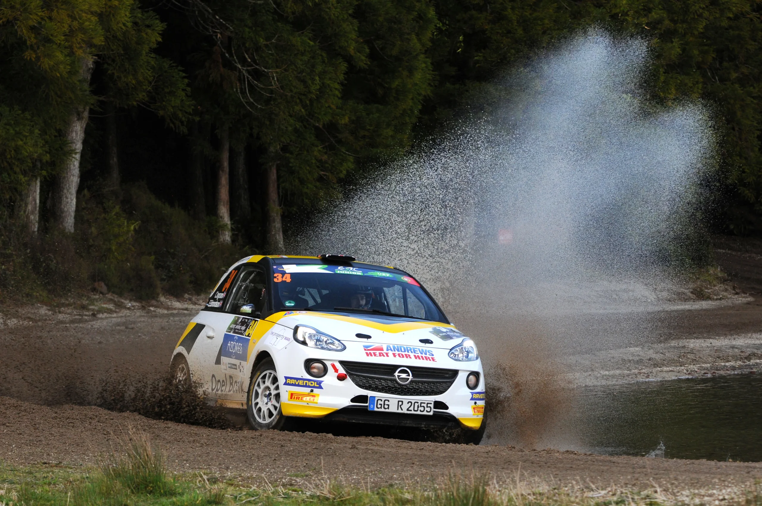 White and yellow Opel Adam R2 rally car carrying number 34 in the ERC Junior category, seen from the front three-quarter as it powers through a water crossing on a forest gravel stage, sending up a broad plume of spray.