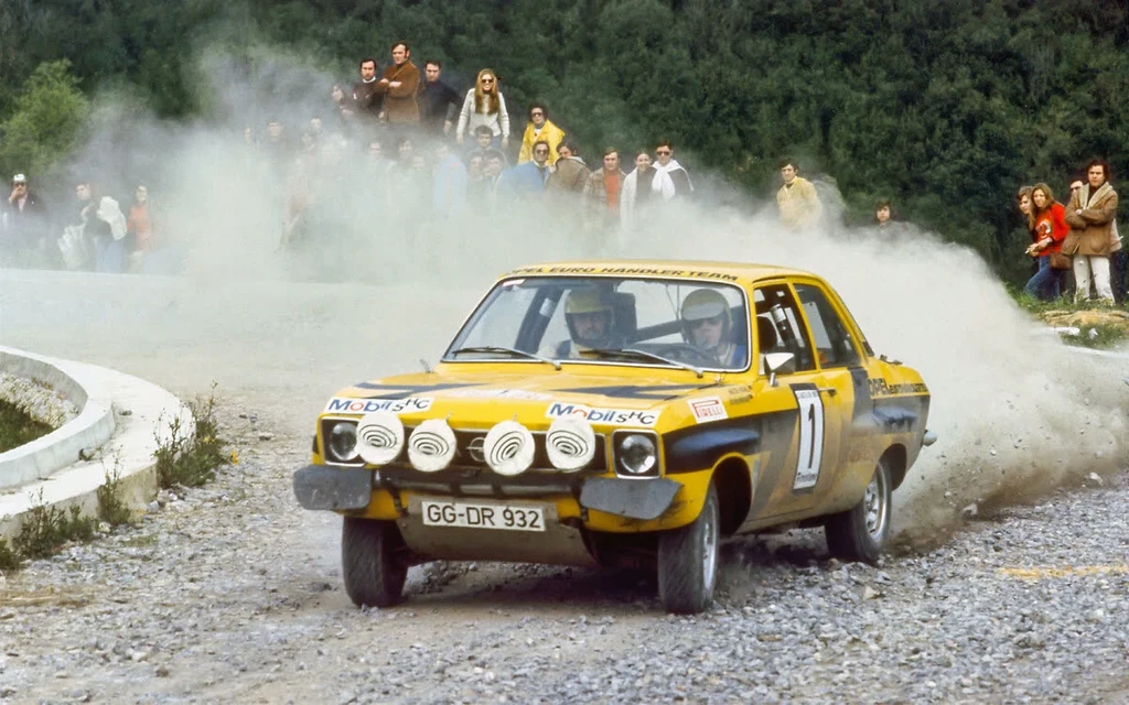 Yellow and black Opel Ascona A rally car with number 1 and four auxiliary lights drifting on a gravel road, kicking up dust in front of spectators.