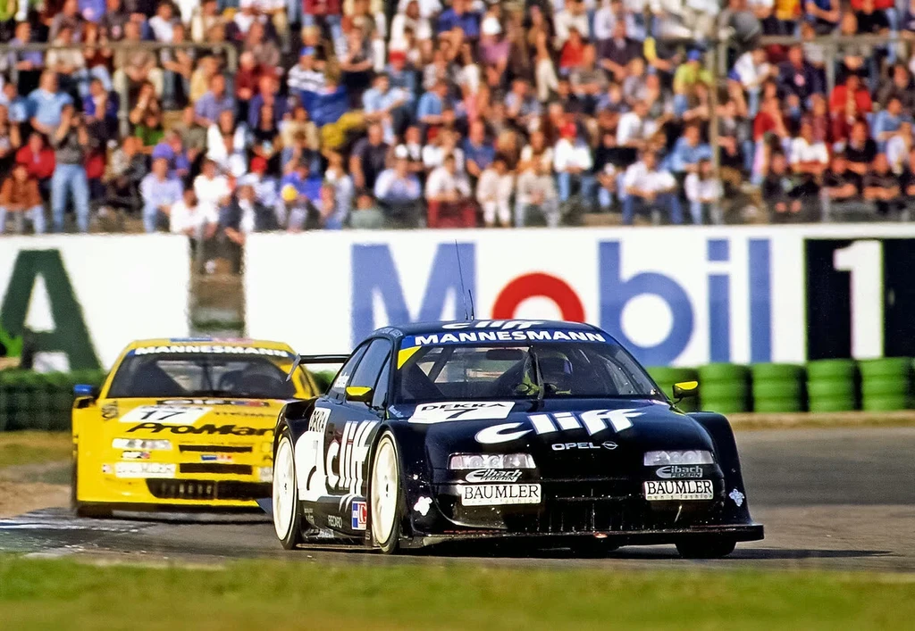 Two Opel Calibra touring cars racing nose to tail on a circuit in the 1990s, the leading car in dark blue Cliff livery ahead of a yellow one, with packed grandstands and trackside advertising behind them.