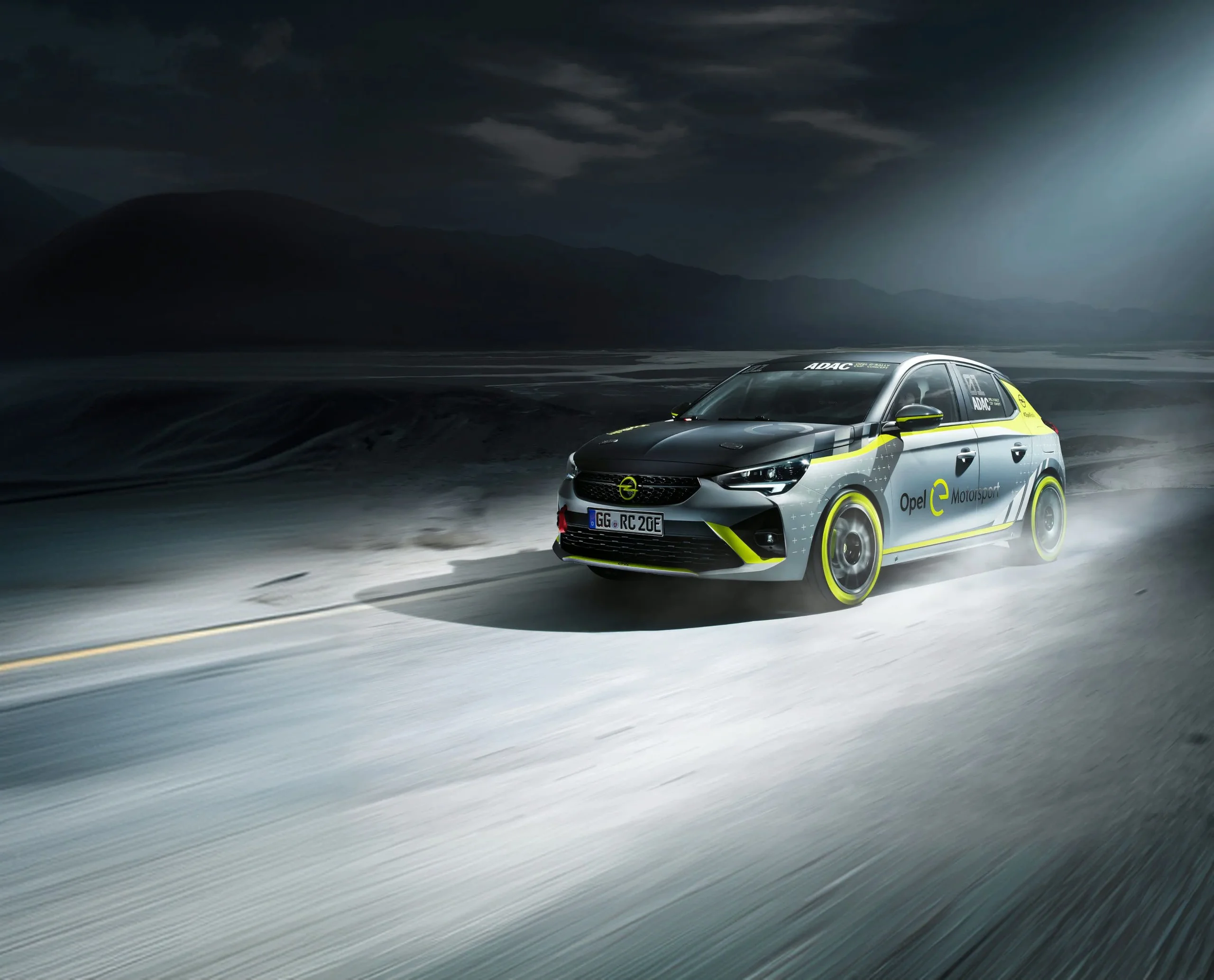 Grey and black Opel Corsa-e rally car with neon yellow accents and yellow-rimmed wheels, seen from the front three-quarter as it drives along a road at dusk, lit against a dark mountain landscape. "Opel e Motorsport" lettering on the door.