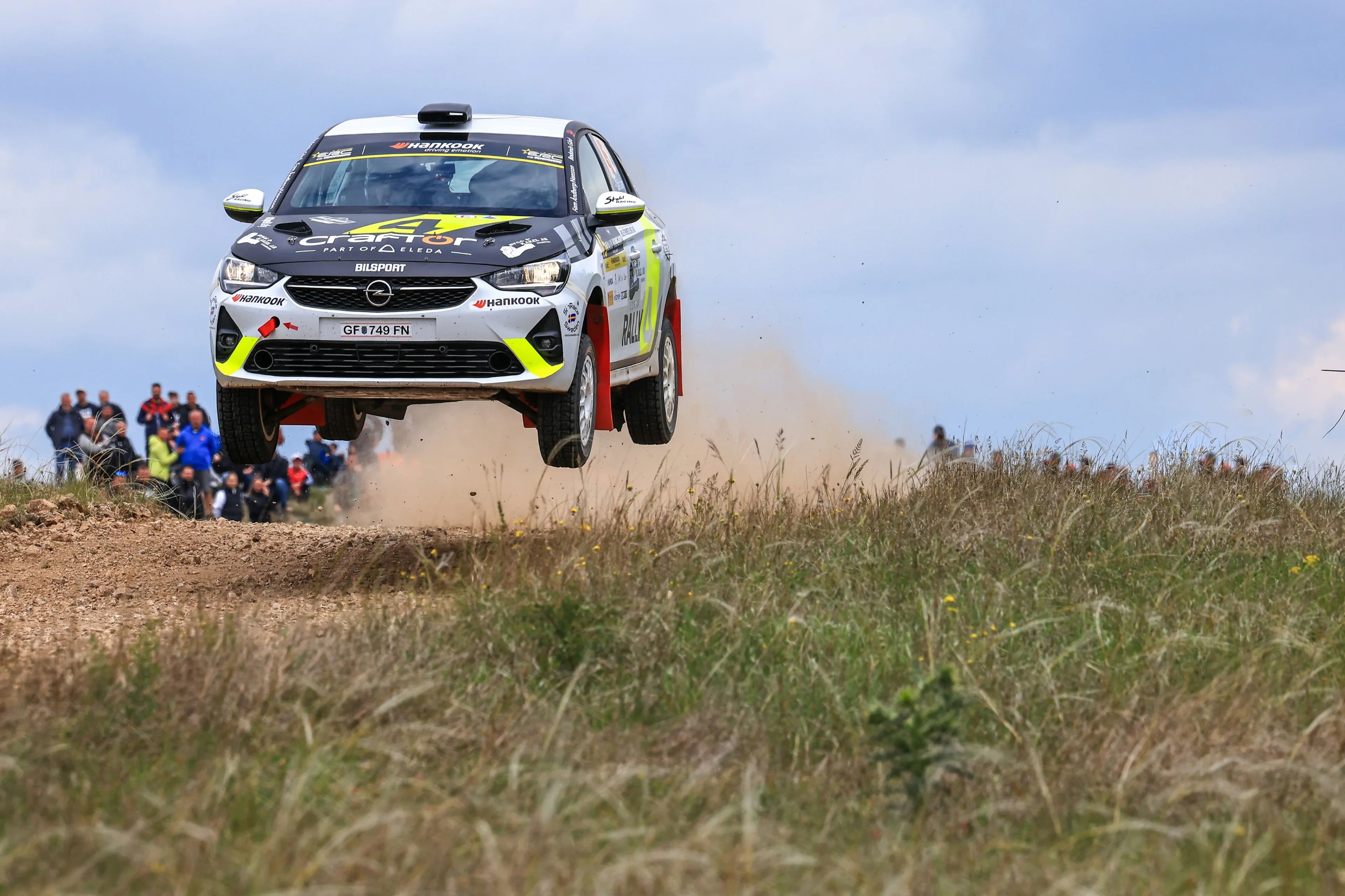 White Opel Corsa Rally car with black bonnet and yellow accents, airborne over a gravel crest with all four wheels off the ground, trailing dust, with spectators lining the bank to the left.