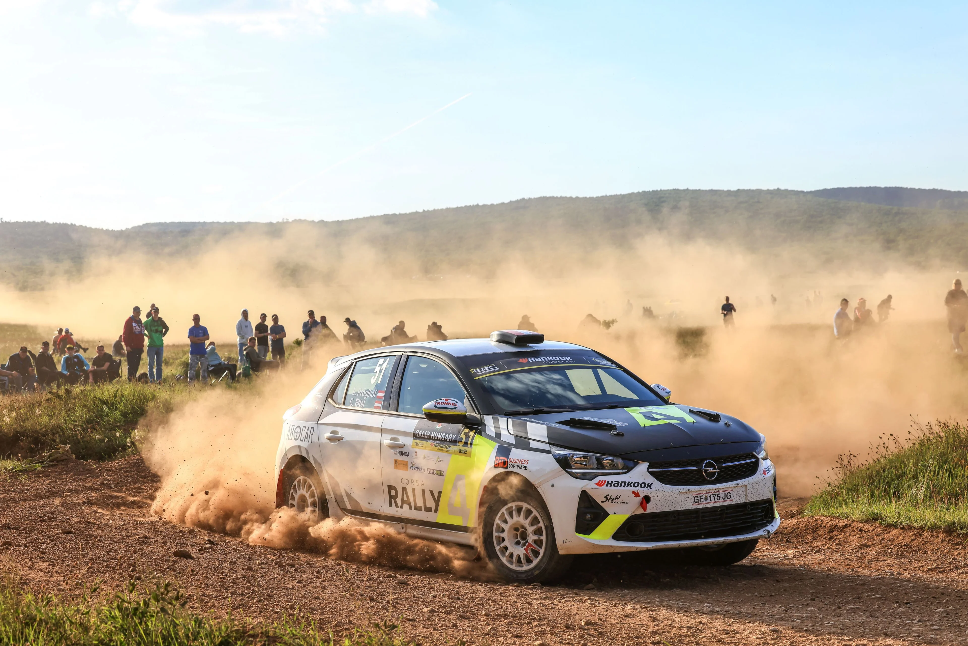 White Opel Corsa Rally car with a black bonnet and yellow accents carrying number 51 at Rally Hungary, sliding through a gravel corner in a thick cloud of dust, with spectators watching from the field behind.