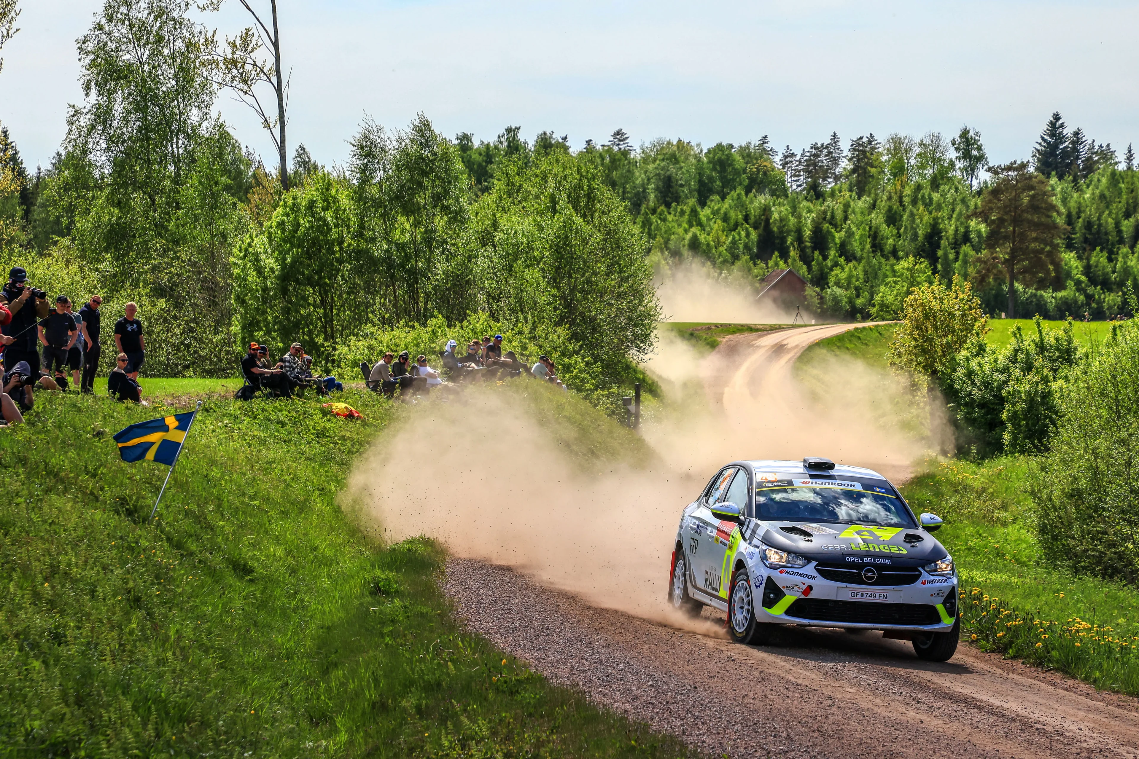White, black and neon yellow Opel Corsa Rally car in Opel Belgium livery on a gravel stage through green countryside, trailing a long cloud of dust, with spectators and a Swedish flag on the bank to the left.