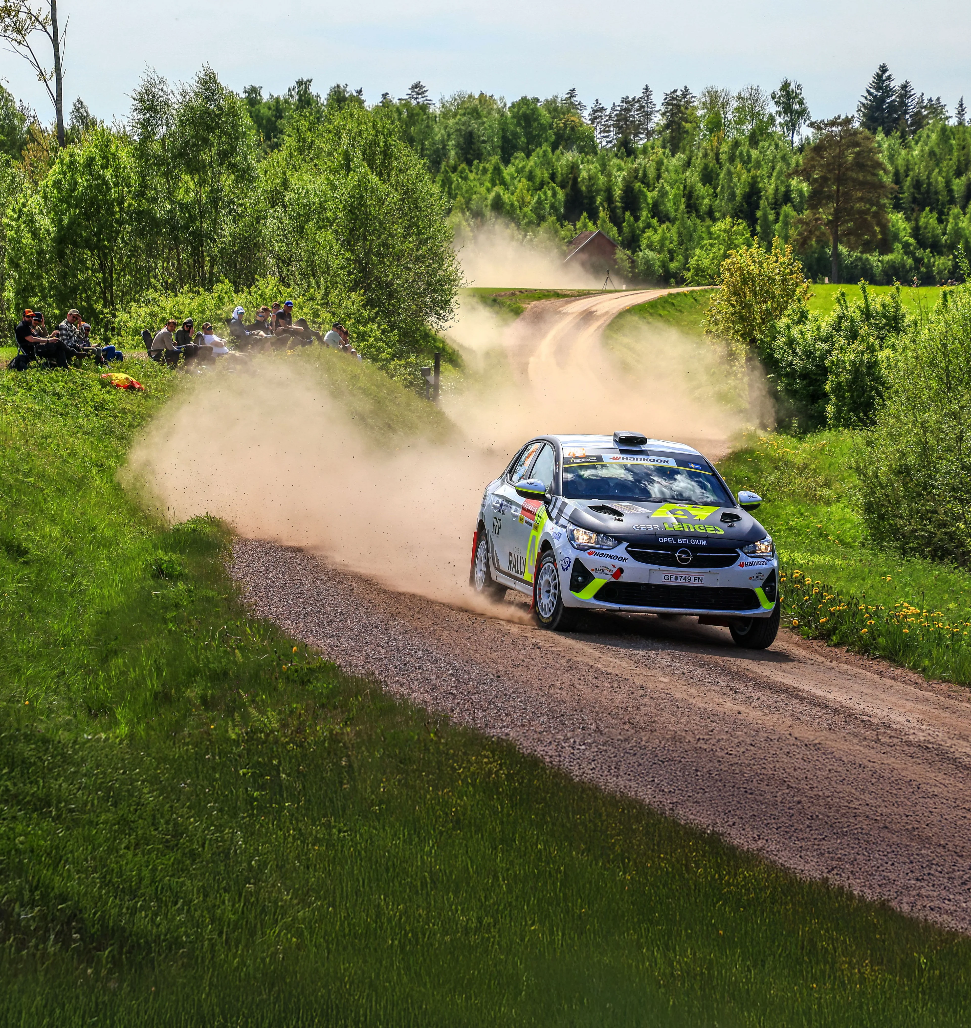 White, black and neon yellow Opel Corsa Rally car in Opel Belgium livery on a gravel stage through green countryside, trailing a long cloud of dust, with spectators and a Swedish flag on the bank to the left.