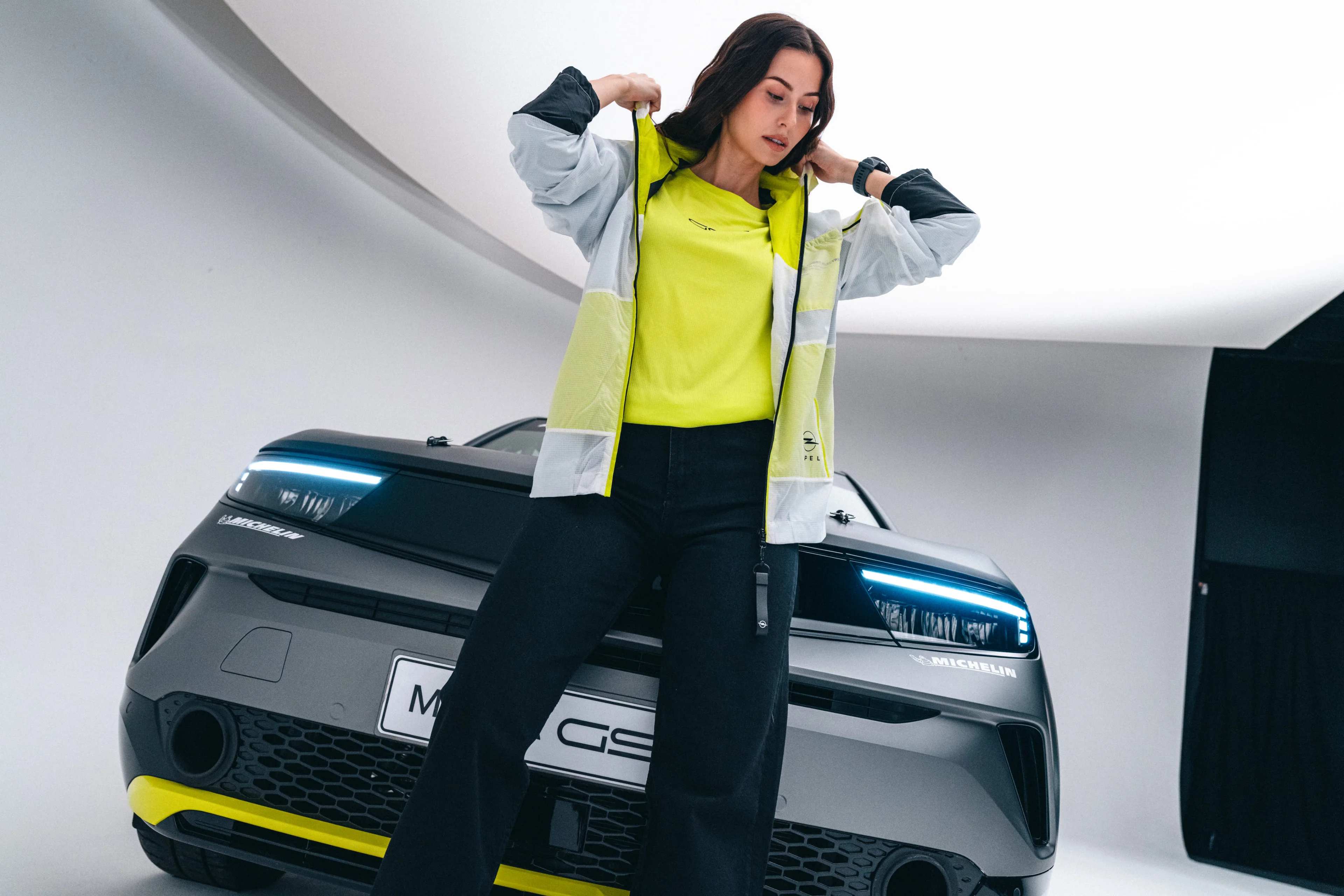 A woman putting on a lightweight white and neon yellow Opel GSE jacket over a yellow GSE T-shirt and dark wide-leg jeans, standing in a studio in front of the Opel Mokka GSE.