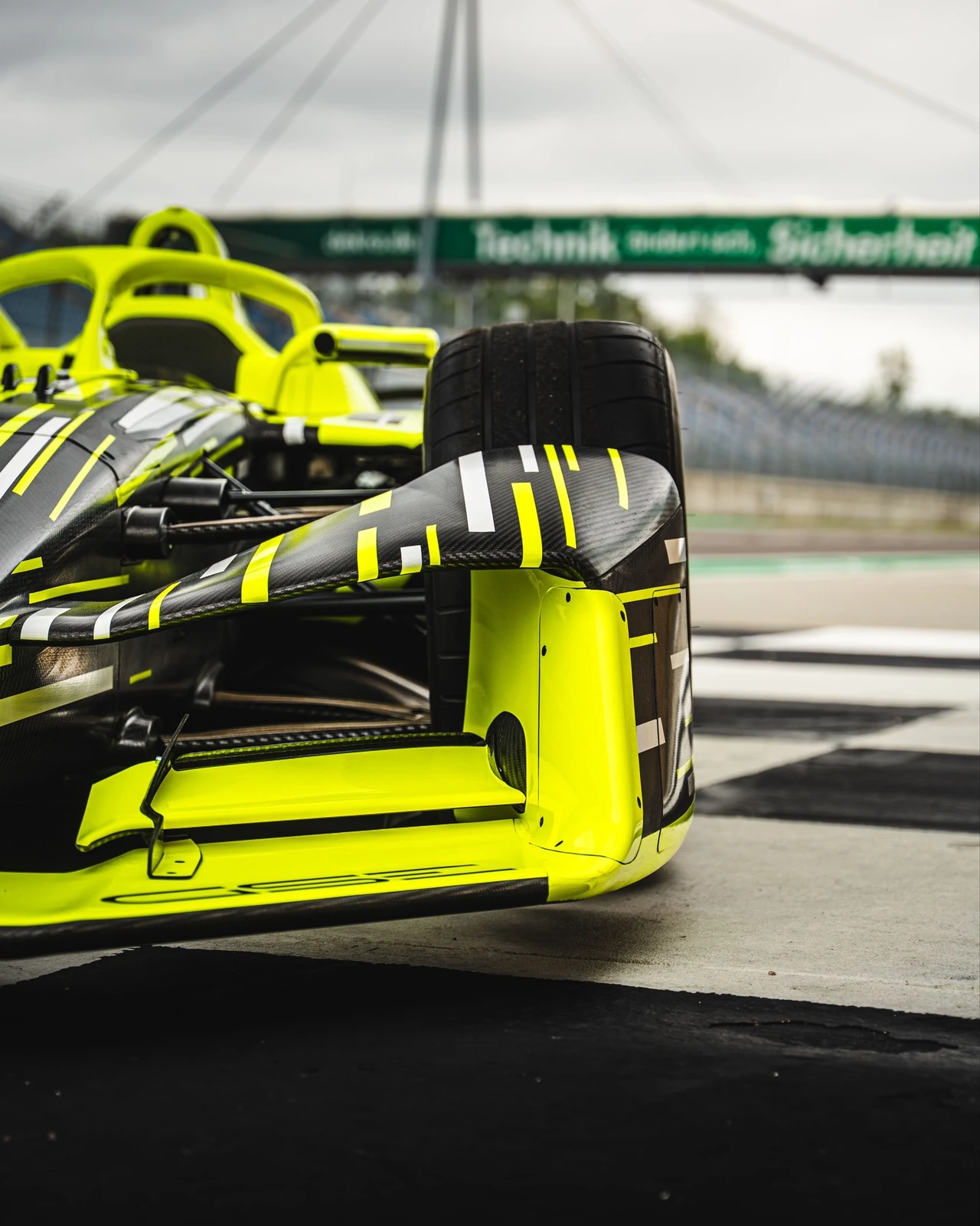 Low close-up of the front wing and wheel of the neon yellow and black Opel GSE Formula E car standing on the chequered start-finish line, with the halo and carbon fibre bodywork behind and a green gantry banner blurred in the distance.