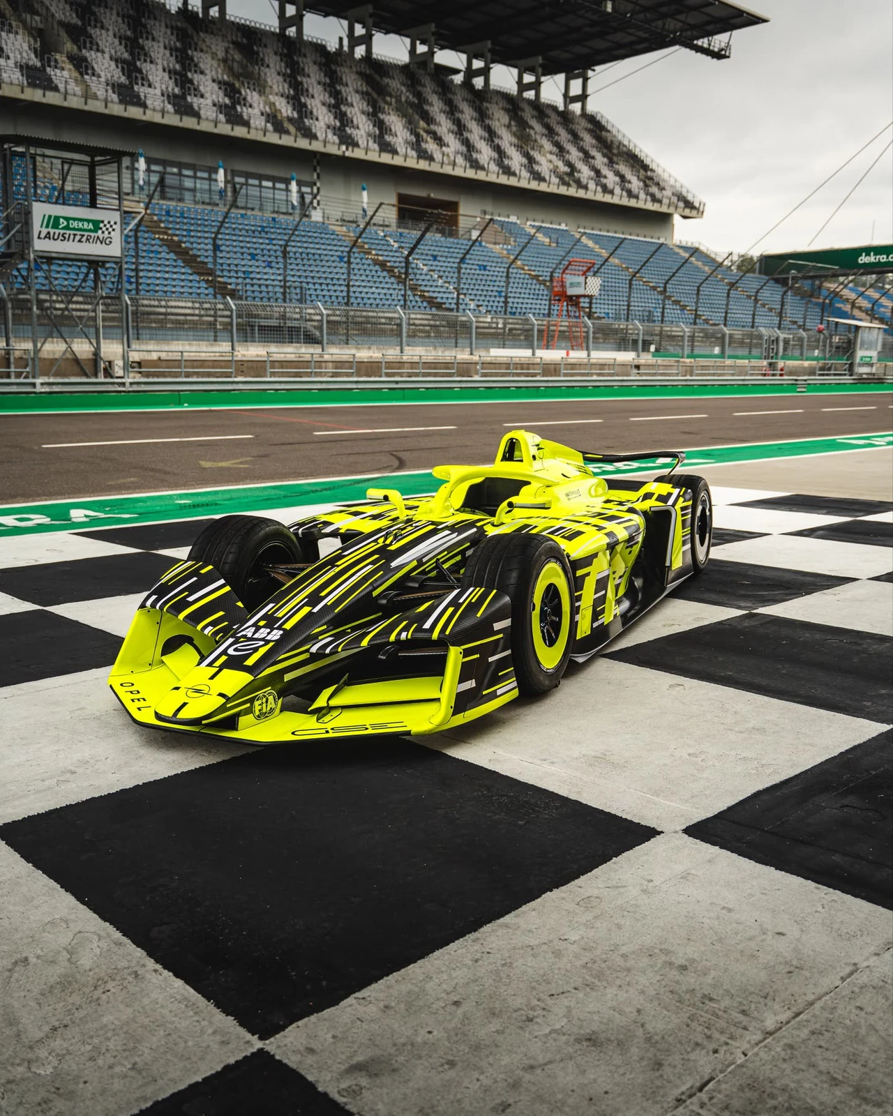 Neon yellow and black Opel GSE Formula E car standing on the chequered start-finish line at the DEKRA Lausitzring, seen from the front three-quarter with empty blue grandstands behind it.