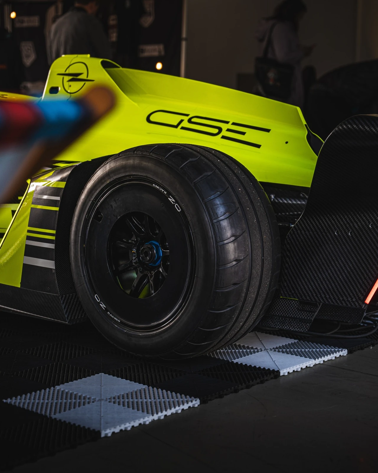 Detail view of the rear wheel of the neon yellow Opel GSE Formula E car in a dimly lit garage, with the GSE lettering and Opel Blitz on the engine cover above the OZ Racing wheel and carbon fibre bodywork.
