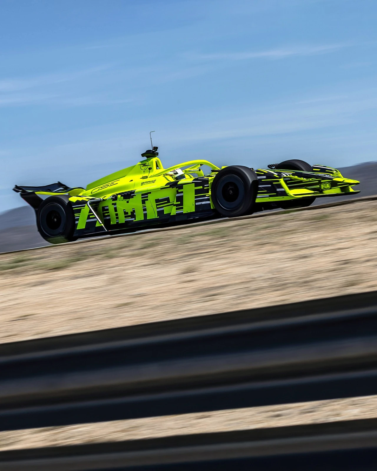 Neon yellow and black Opel GSE Formula E car seen in side profile at speed on a circuit, the driver visible in the cockpit and the barrier and dry hillside blurred by a panning shot.