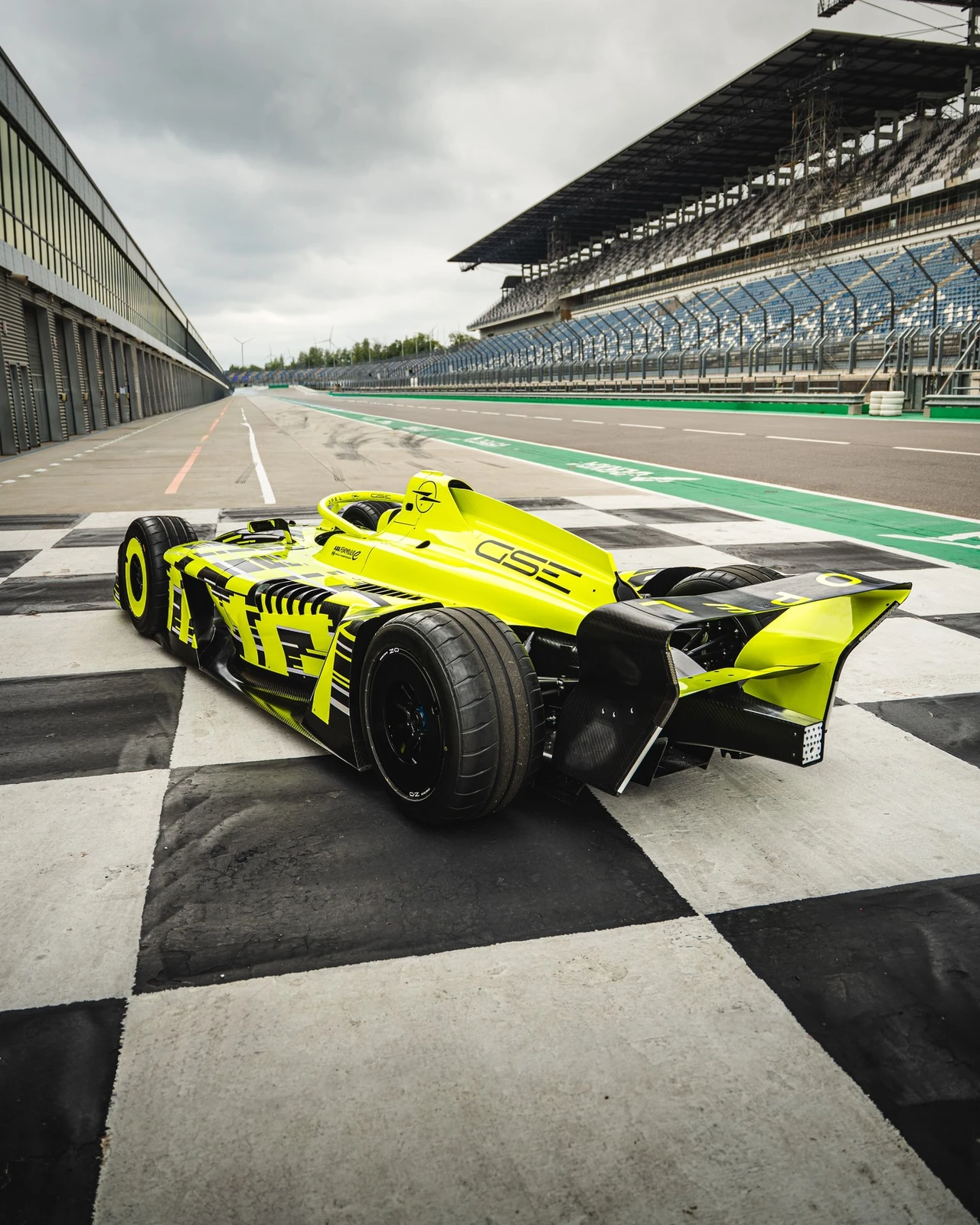 Neon yellow and black Opel GSE Formula E car standing on the chequered start-finish line of a race circuit, seen low from the rear three-quarter, with the pit building on one side and empty grandstands under a grey sky on the other.