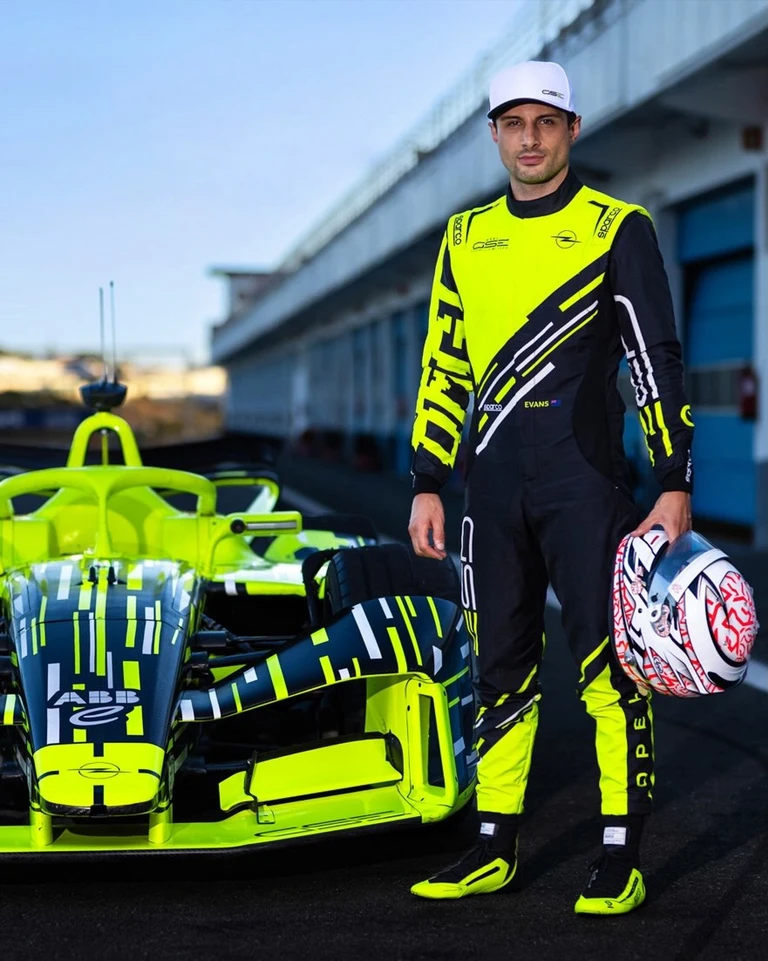Formula E driver in a neon yellow and black Opel GSE race suit and white GSE cap, holding his helmet and standing in the pit lane beside the neon yellow Opel GSE Formula E car.