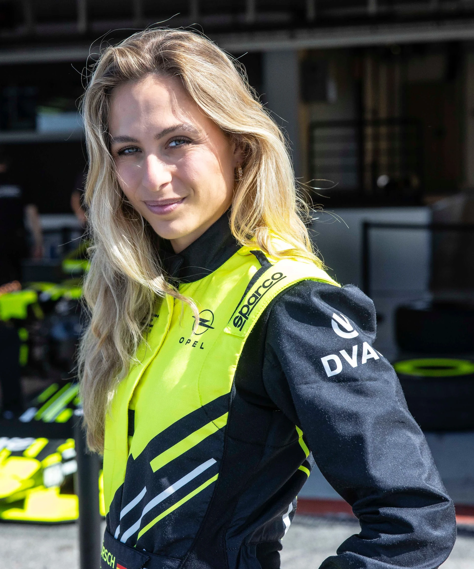A driver in a neon yellow and black Opel GSE race suit standing in the pit lane, with the team working on a single-seater in the garage behind her.