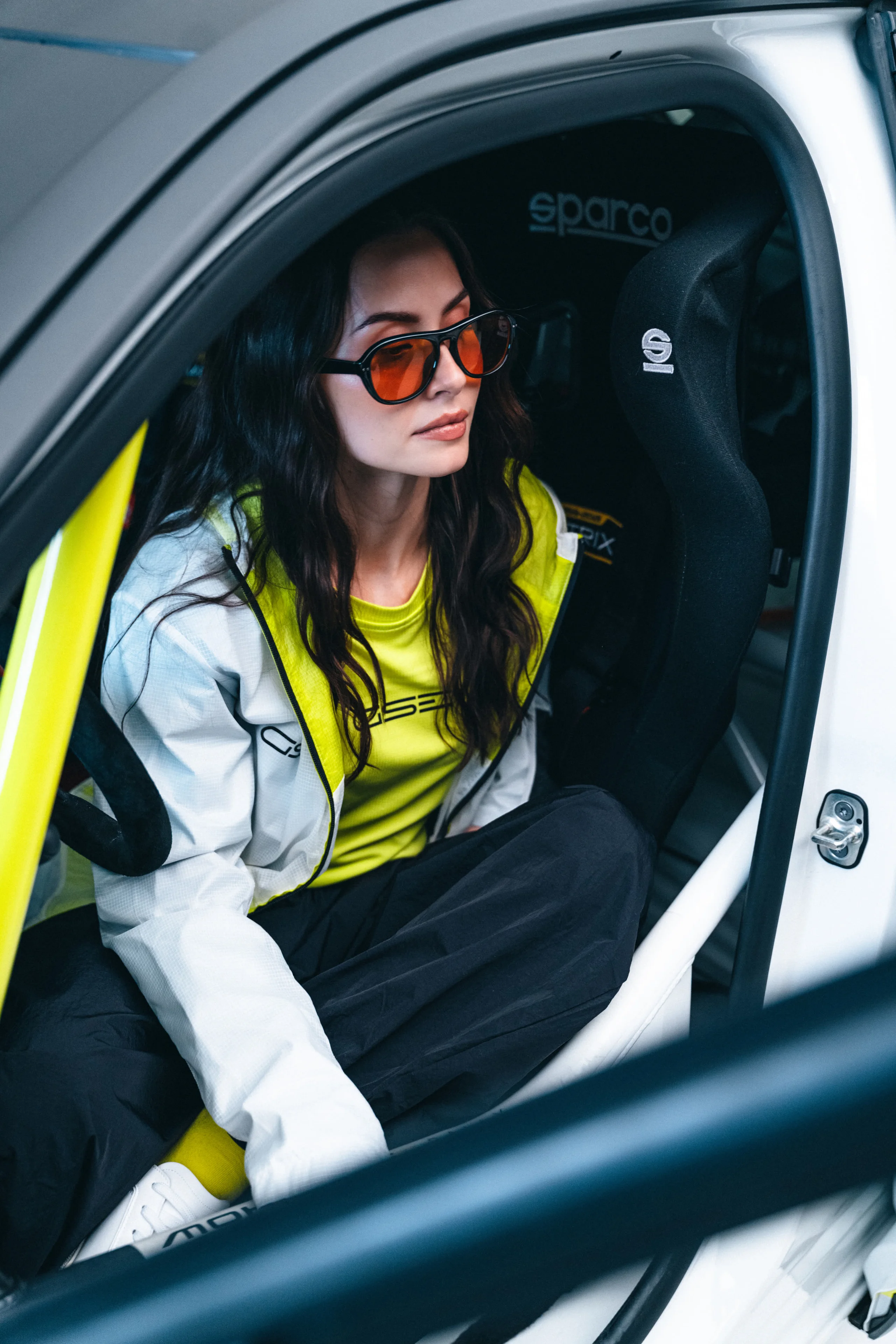 A woman in sunglasses wearing a white and neon yellow Opel GSE zip jacket over a yellow GSE T-shirt, sitting sideways in the open door of a rally car with a black Sparco bucket seat behind her.