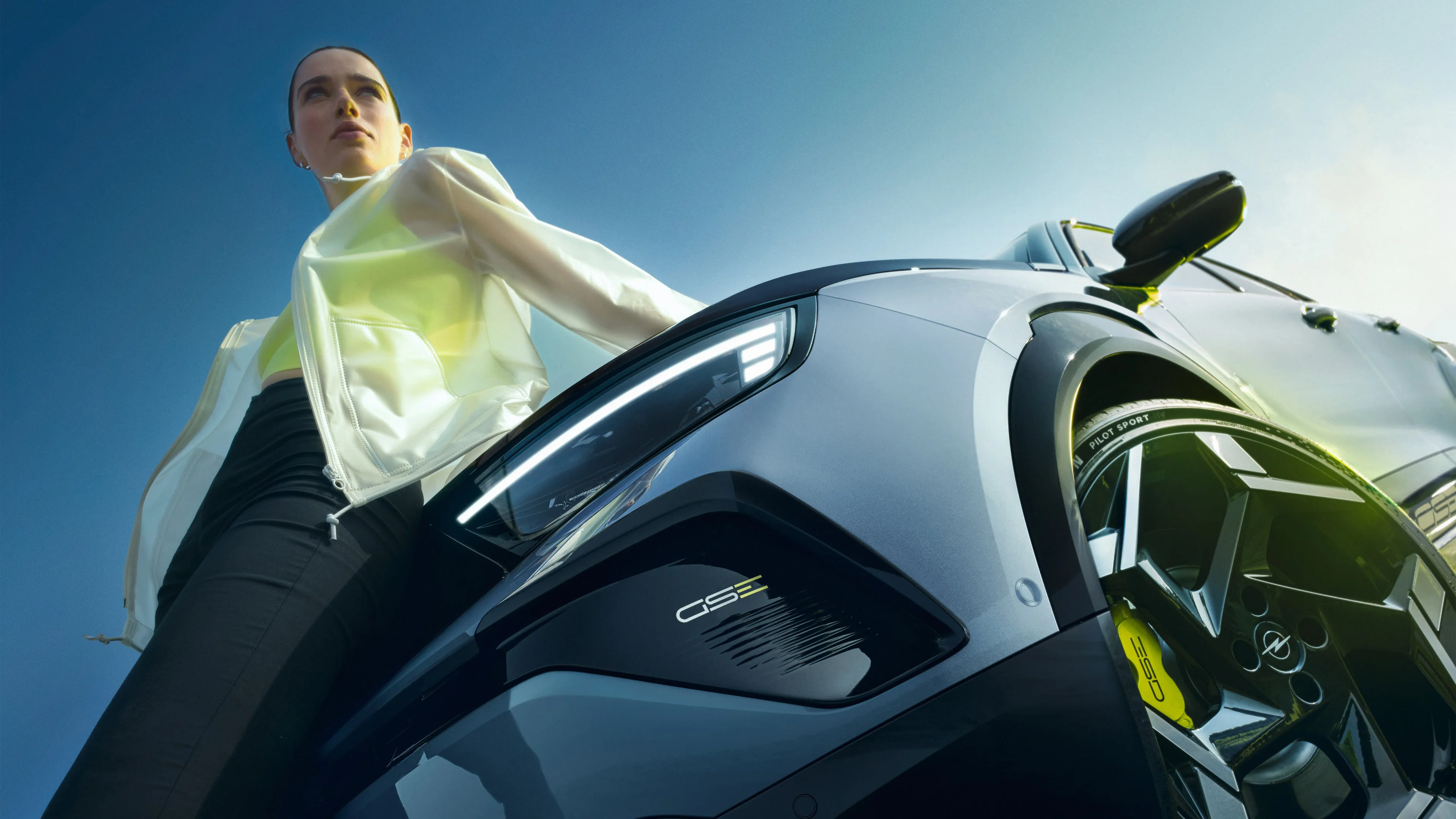 Low-angle shot of a woman in a white and yellow Opel GSE jacket standing beside a silver-grey Opel GSE, with the car's headlight, GSE badge and yellow brake caliper prominent in the foreground against a clear blue sky.