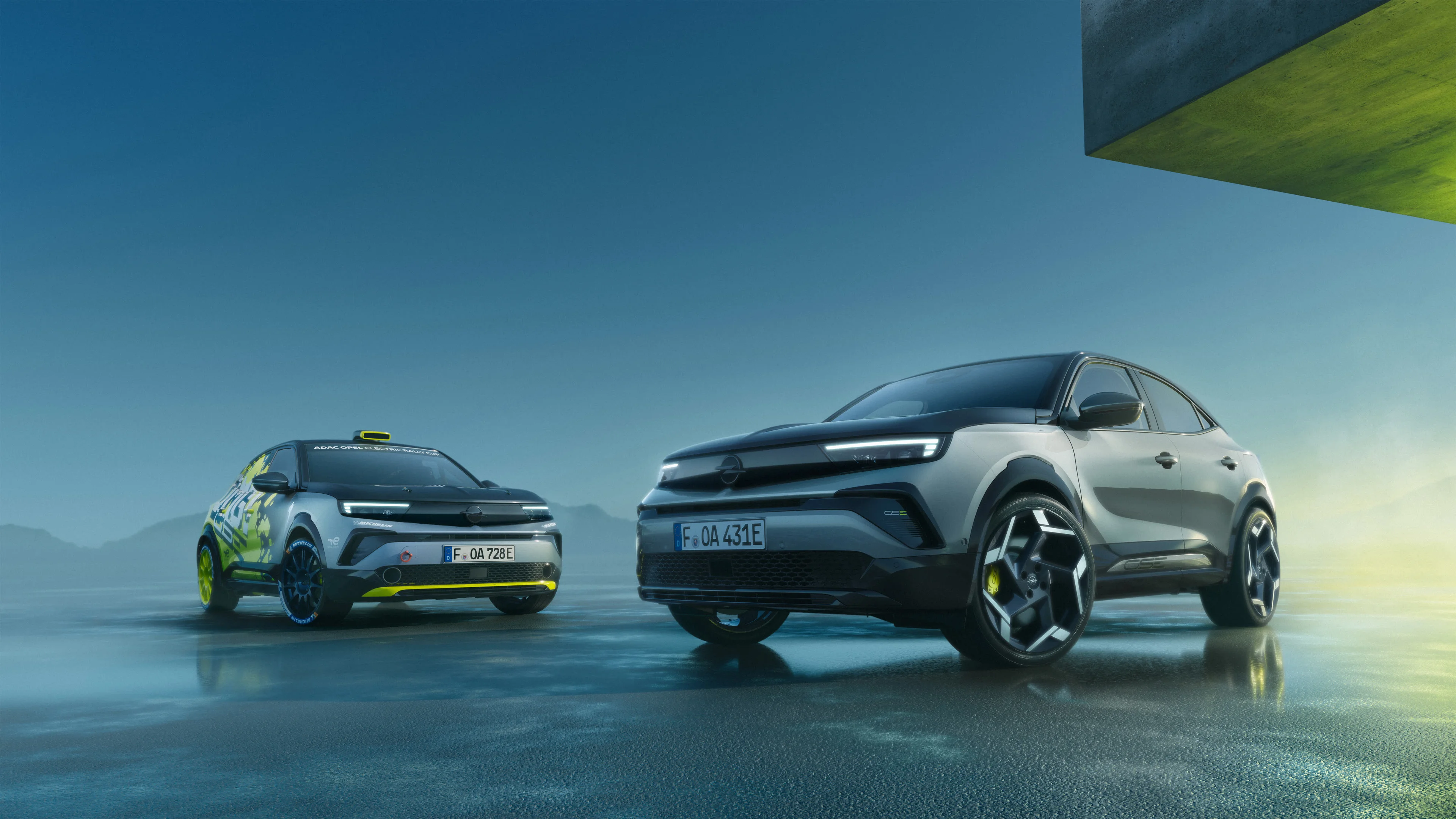 Two Opel Mokka GSE models on a wet reflective surface at dusk, both seen from the front: the silver-grey production version on the right and the yellow and black rally version with roof light on the left.