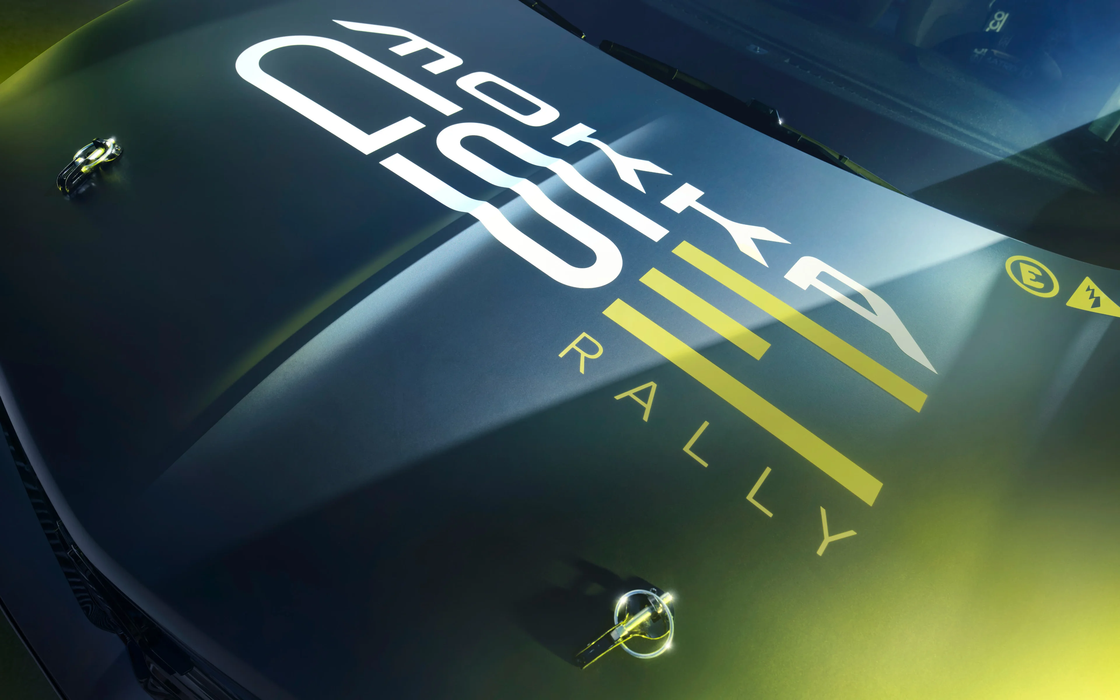 Close-up of the dark bonnet of the Opel Mokka GSE Rally car, showing the white "MOKKA GSE" lettering and yellow "RALLY" graphic, with two bonnet pins and a high-voltage warning sticker.