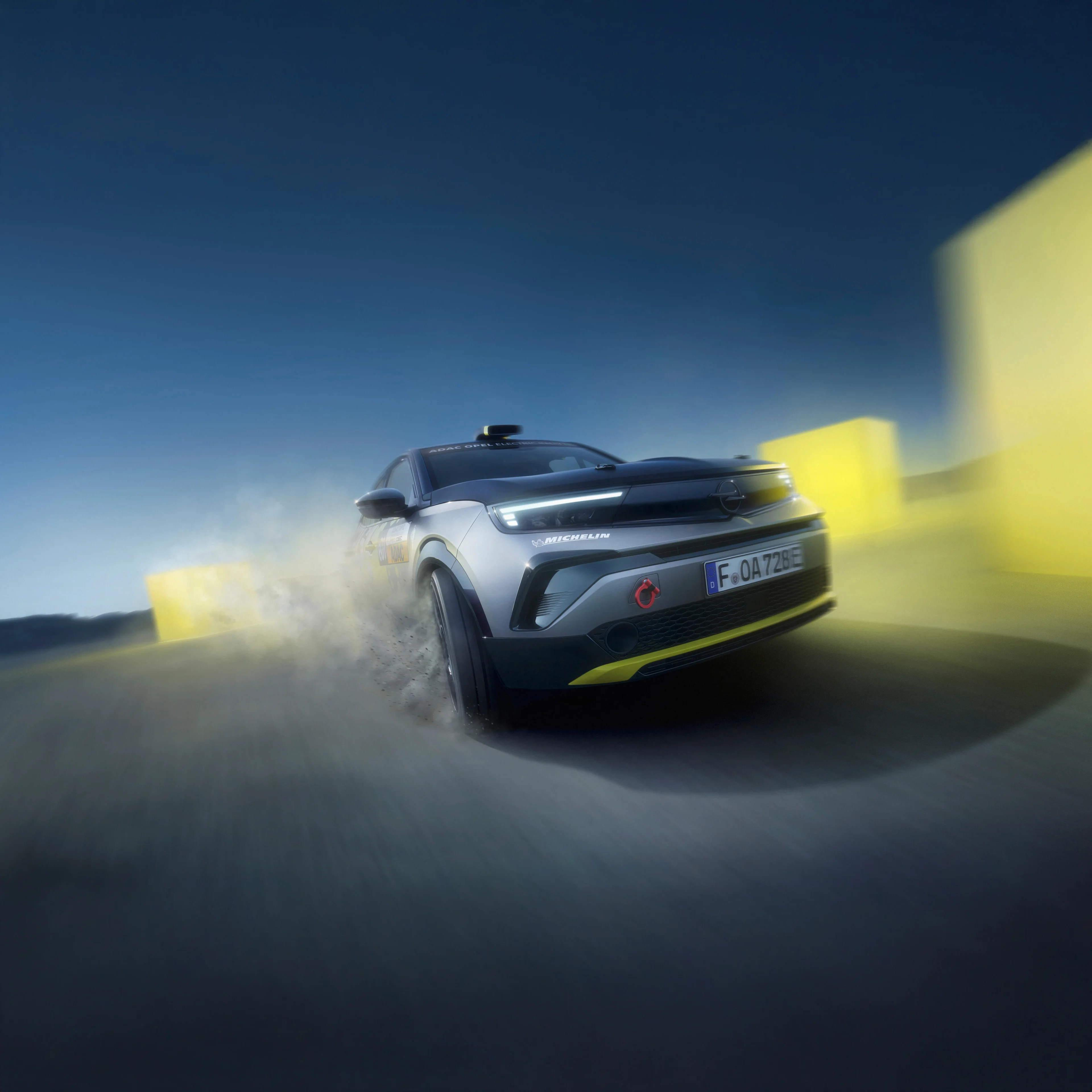 Opel Mokka GSE rally car seen head-on driving towards the camera at dusk, throwing up dust, with blurred yellow blocks marking the course and a deep blue sky behind.