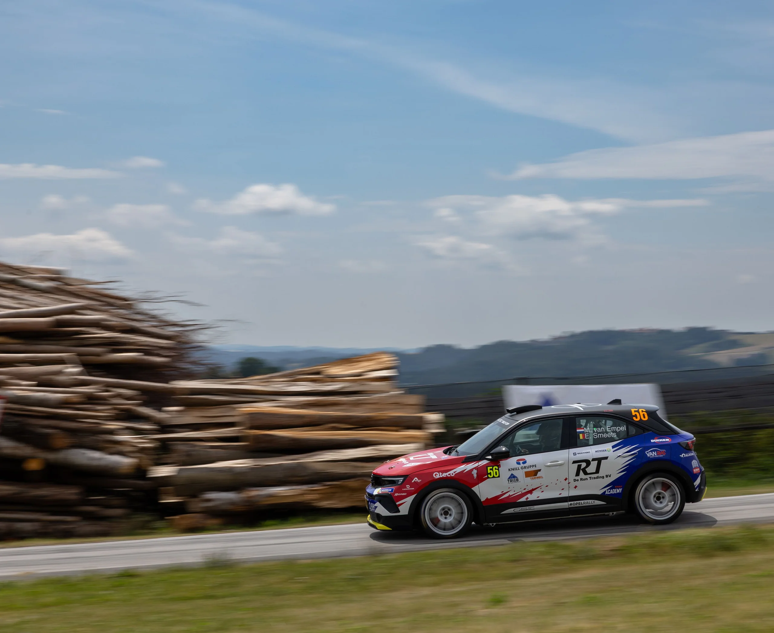 Opel Mokka GSE Rally car in red, white and blue livery carrying number 56, seen in side profile at speed on a tarmac stage past a stack of felled timber, with rolling hills behind and the background blurred by a panning shot.