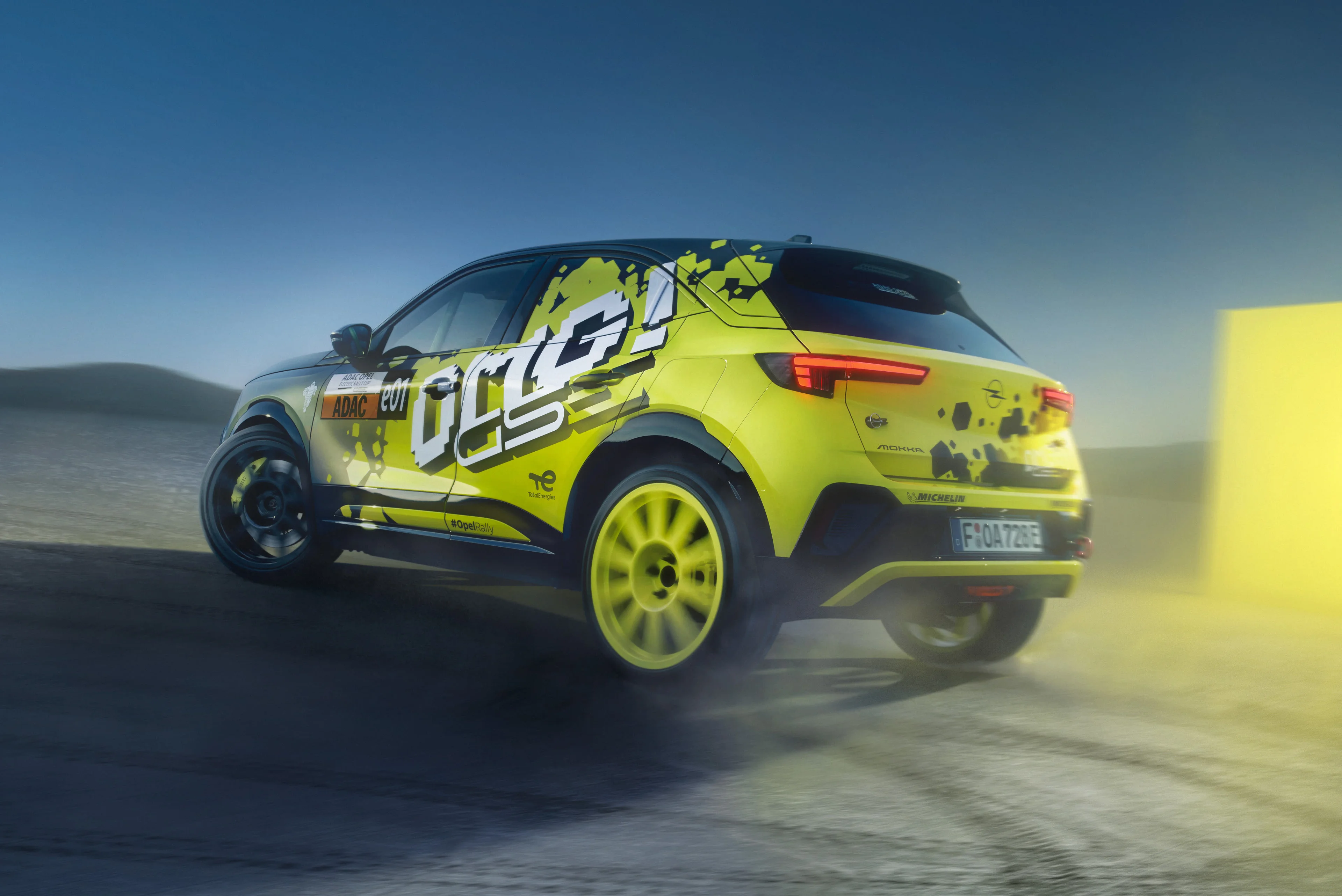 Neon yellow and black Opel Mokka GSE Rally car in "OMG! GSE" livery, seen from the rear three-quarter at dusk with its tail lights lit, kicking up dust on a gravel surface with a yellow course marker glowing behind.