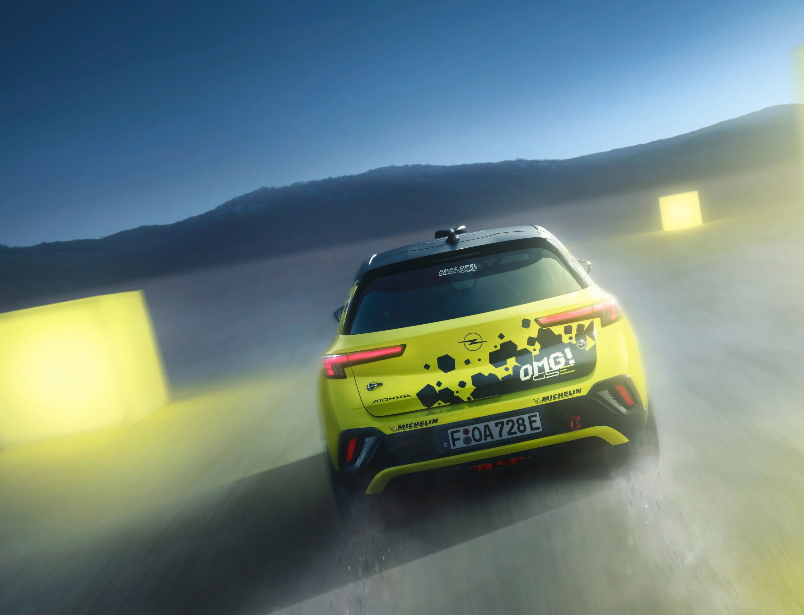 Neon yellow Opel Mokka GSE Rally car seen straight from behind driving away at dusk, tail lights lit, with yellow course markers blurred past on either side and a dark ridge on the horizon.