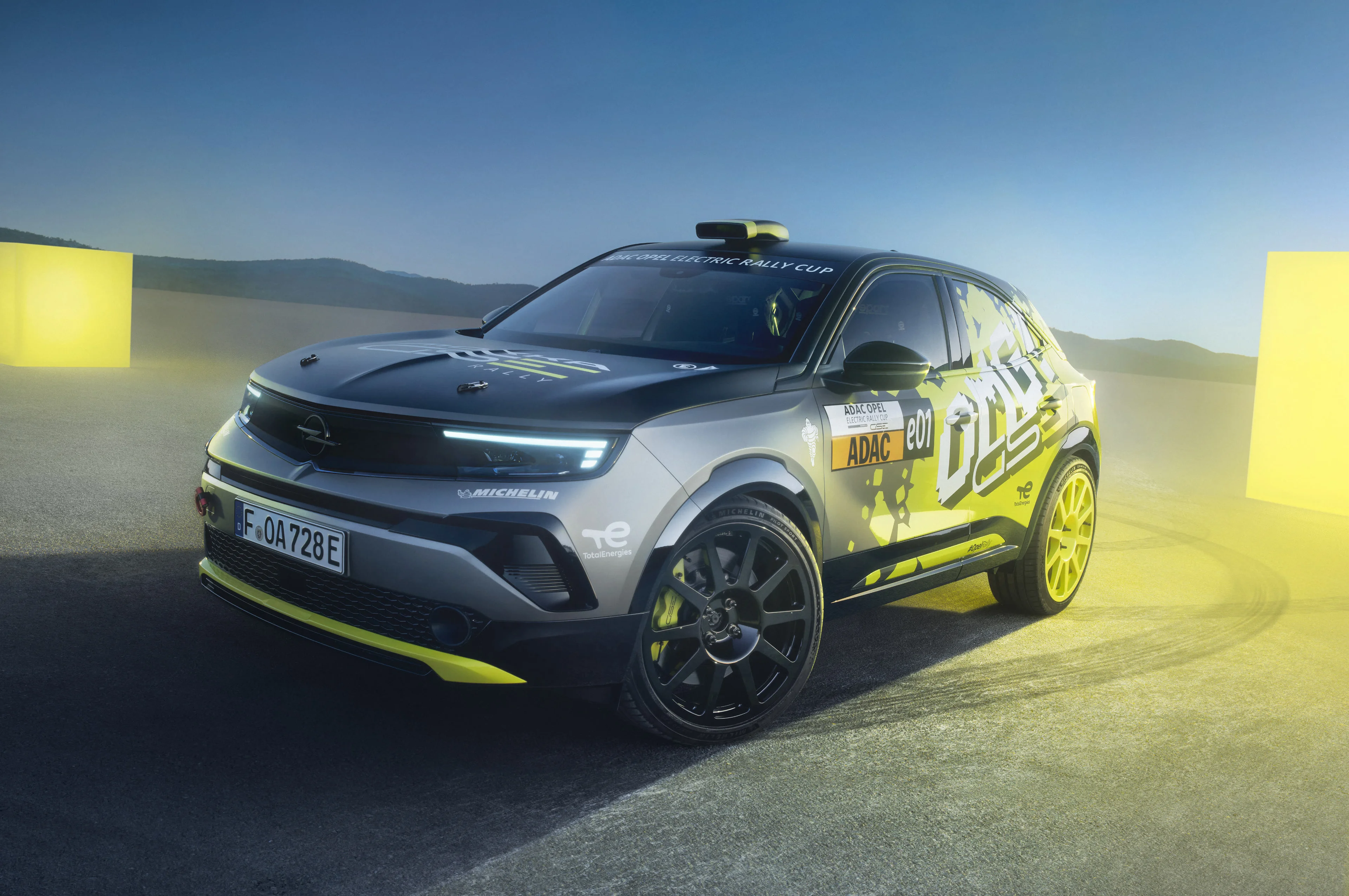 Opel Mokka GSE Rally car in grey with yellow and black "OMG GSE" graphics, standing still on tarmac at dusk between two glowing yellow course markers, seen from the front three-quarter with its daytime running lights on and Michelin and TotalEnergies decals on the front wing.