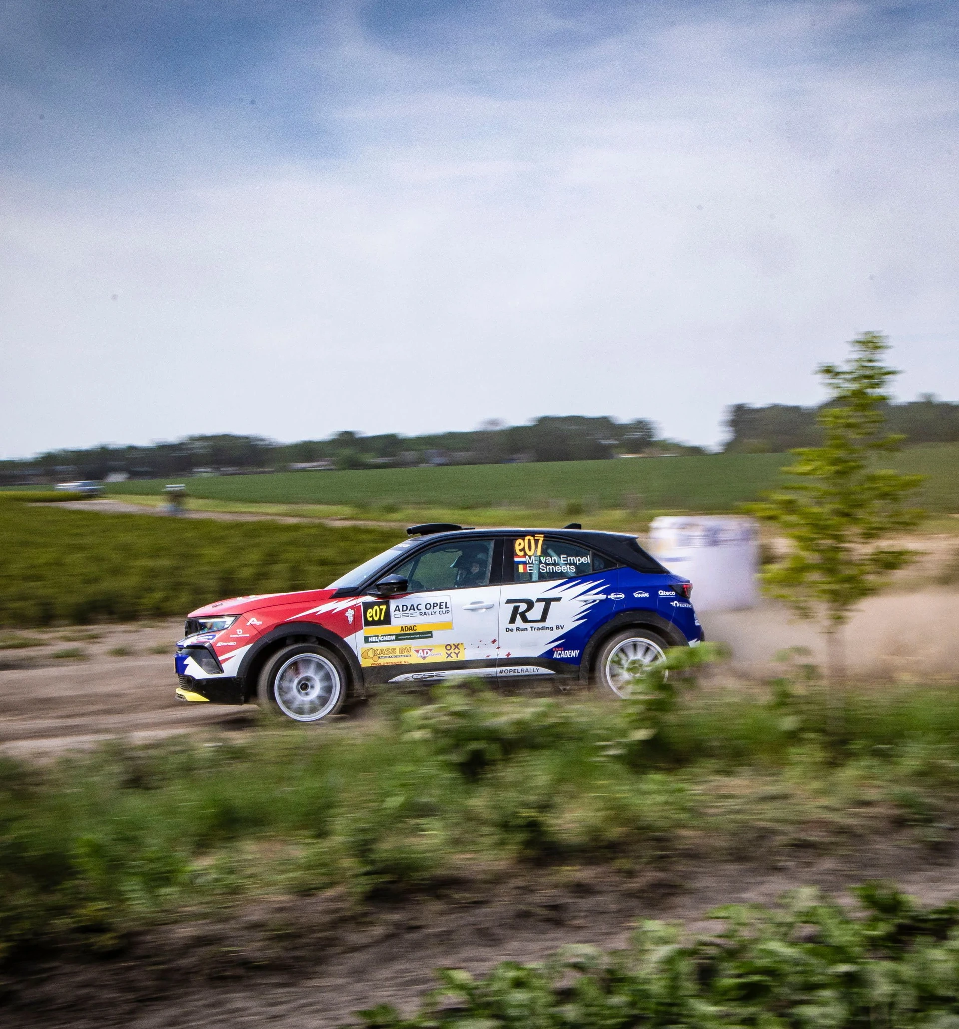 Opel Mokka GSE Rally car in red, white and blue livery carrying number e07 in the ADAC Opel GSE Rally Cup, seen in side profile on a gravel stage through flat farmland, trailing dust with the background blurred by a panning shot.