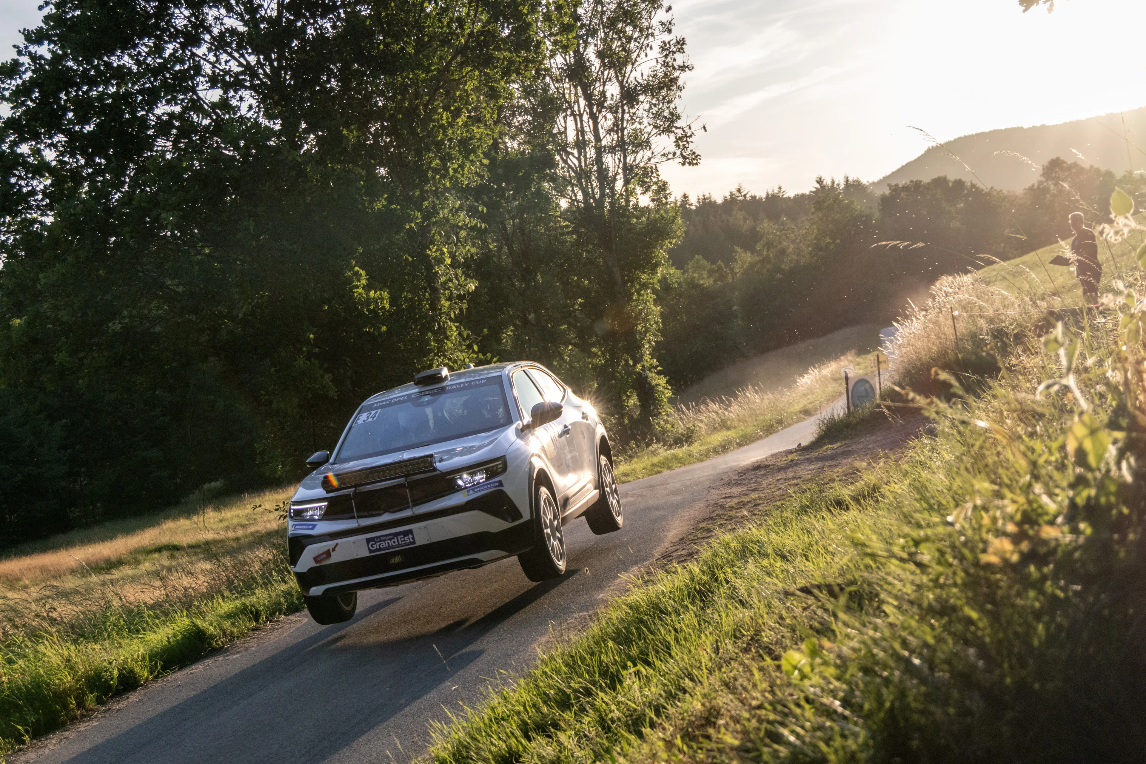 White Opel Mokka GSE Rally car carrying number E 34 in the ADAC Opel GSE Rally Cup, airborne over a crest on a narrow tarmac stage in low evening sunlight, with wooded hills behind.