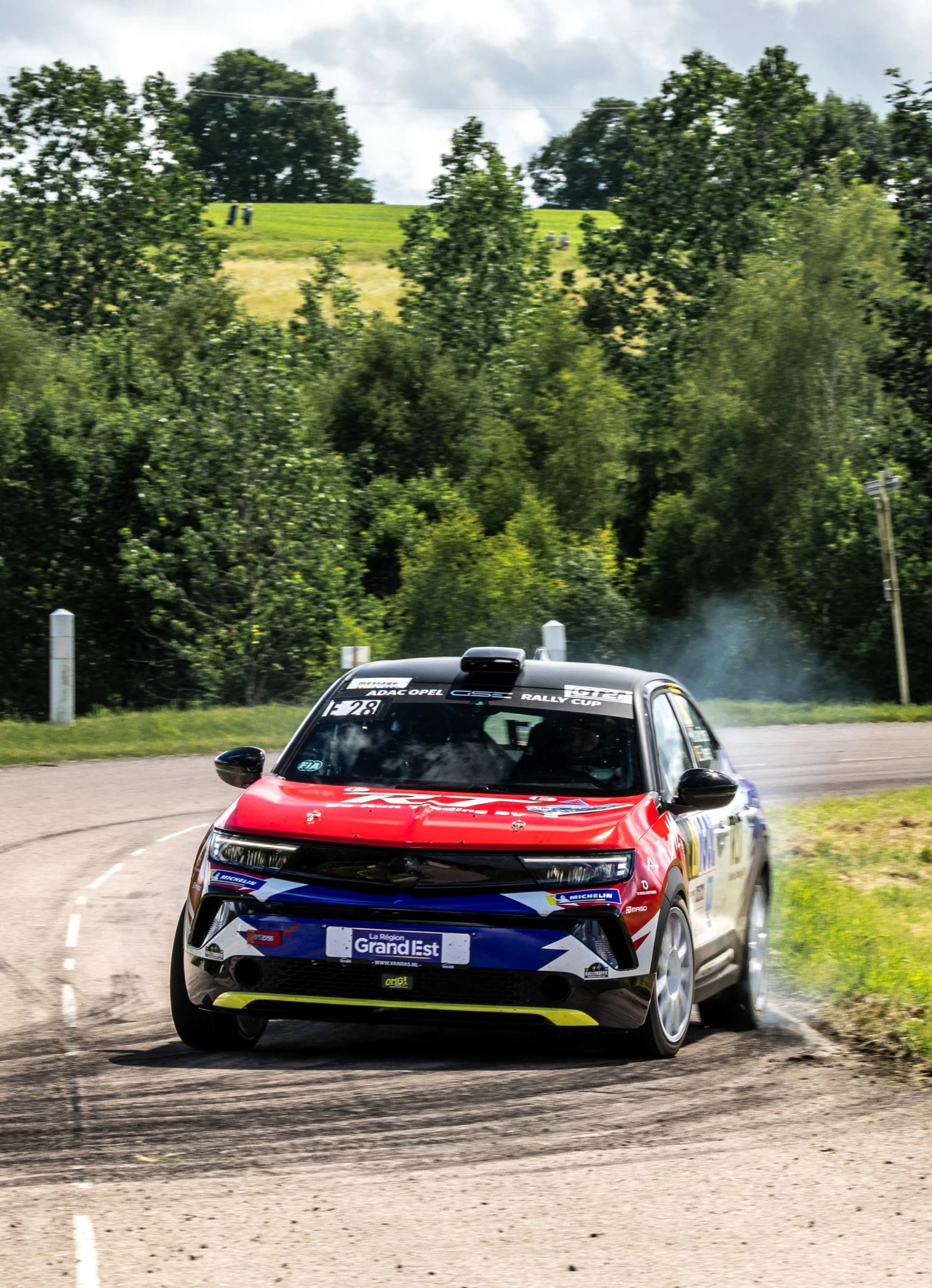 Opel Mokka GSE Rally car in red, white and blue livery carrying number 28 in the ADAC Opel GSE Rally Cup, cornering on a narrow tarmac stage through green farmland with spectators on the hillside behind.