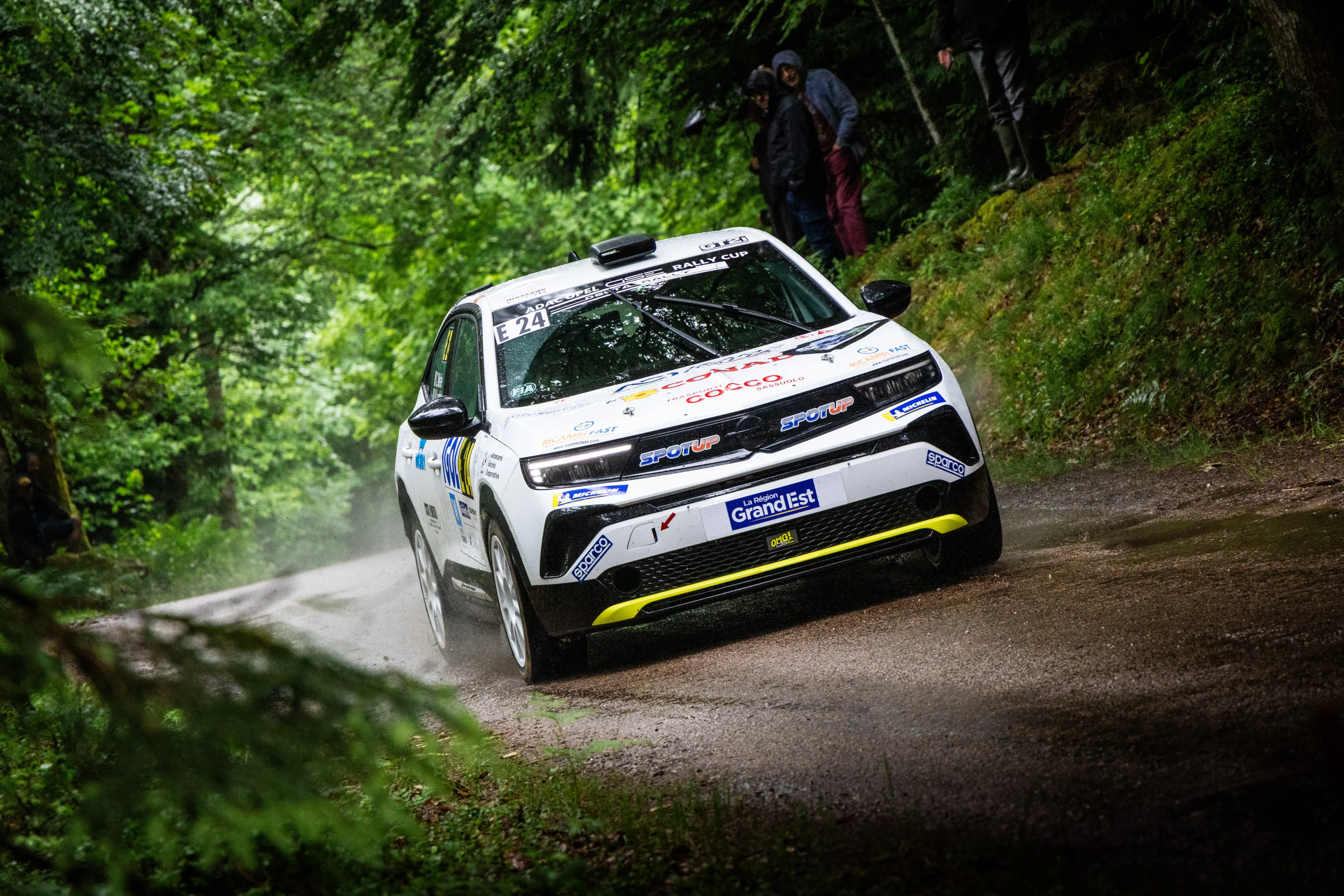 White Opel Mokka GSE Rally car with yellow front splitter carrying number E 24 in the ADAC Opel GSE Rally Cup, cornering on a wet gravel stage through dense green forest, with a few spectators on the bank above.
