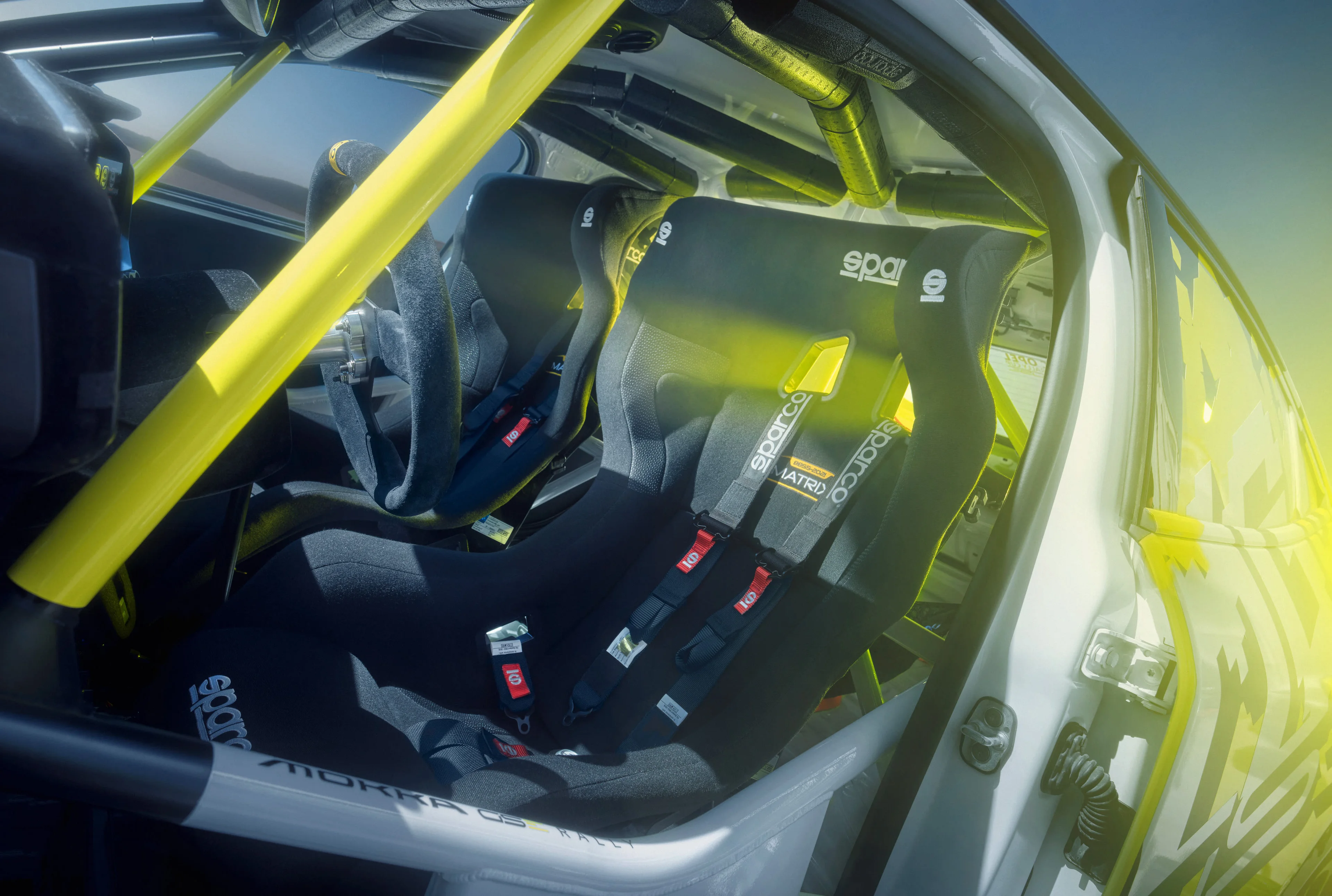 Interior of the Opel Mokka GSE rally car seen through the open door, with two black Sparco bucket seats, four-point harnesses and a yellow roll cage.