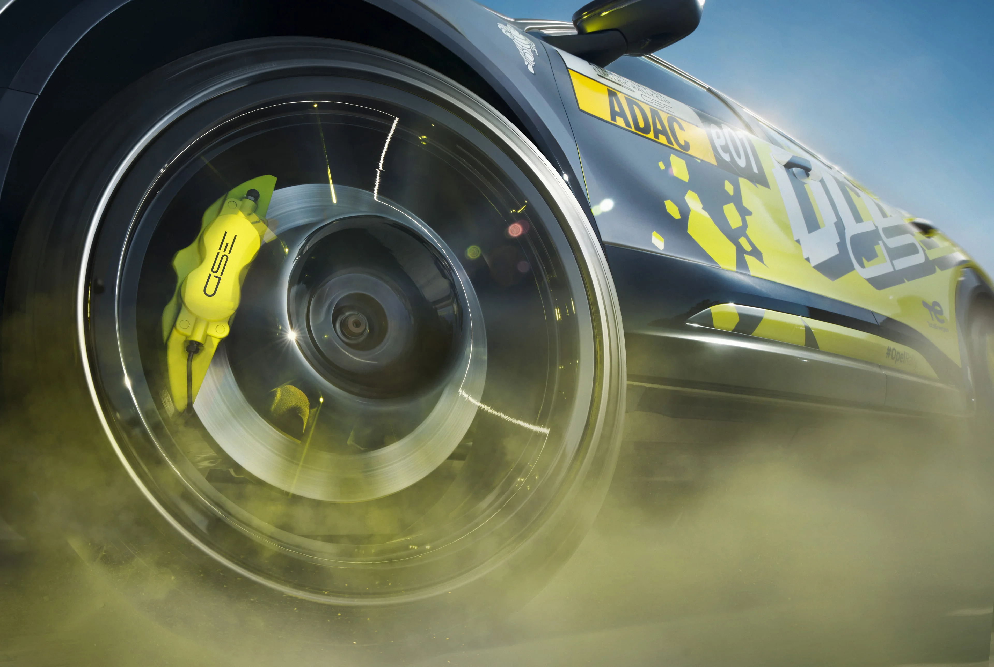 Close-up of a spinning front wheel on the Opel Mokka GSE Rally car, with the yellow GSE brake caliper and brake disc visible through the blurred spokes and dust rising from the tyre.