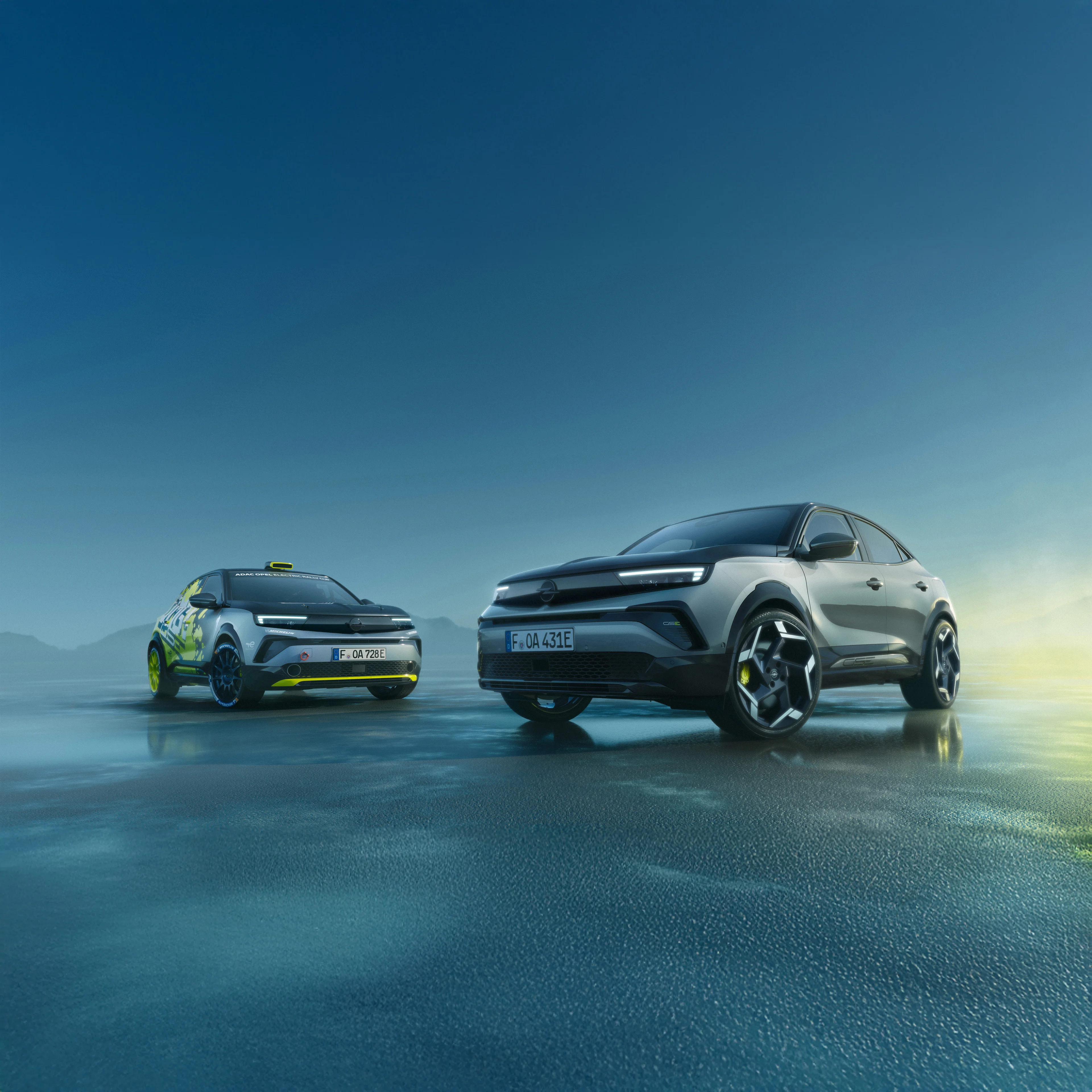 Two Opel Mokka GSE models side by side on a wet reflective surface at dusk: the silver-grey production version in the foreground and the yellow and black rally version with roof light behind it, both shown from the front.