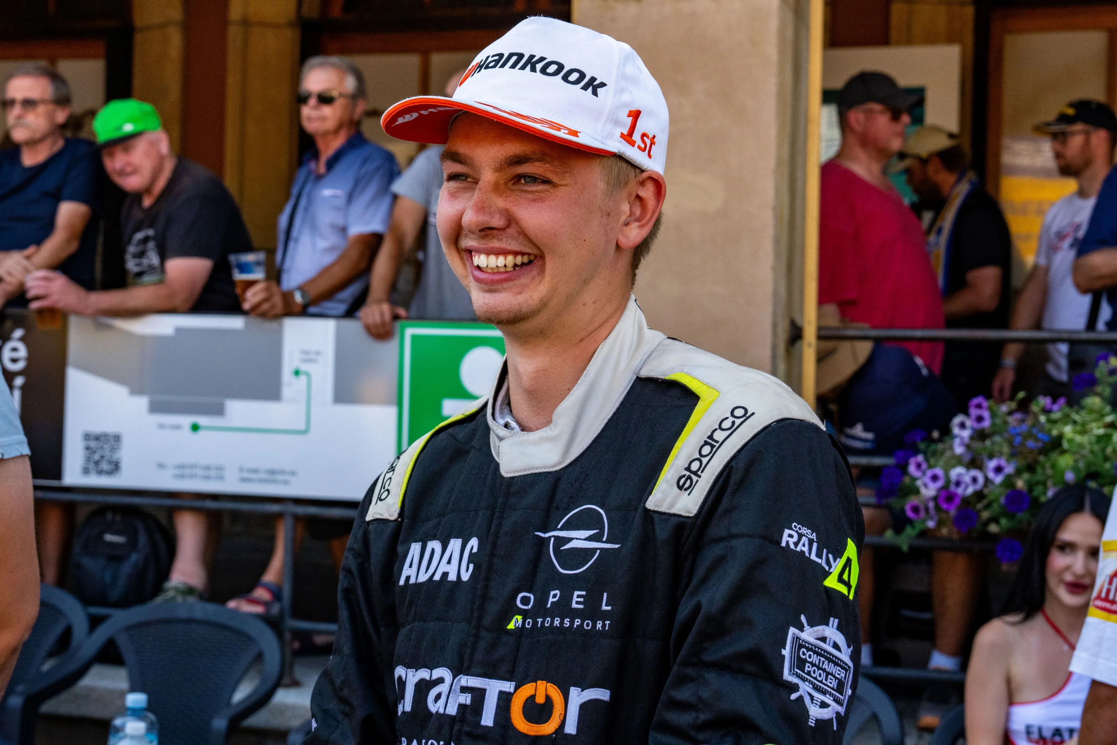 A young driver in a black Opel Motorsport and ADAC racing suit, smiling and wearing a white winner's cap marked "1st", with spectators and a rally finish area behind him.