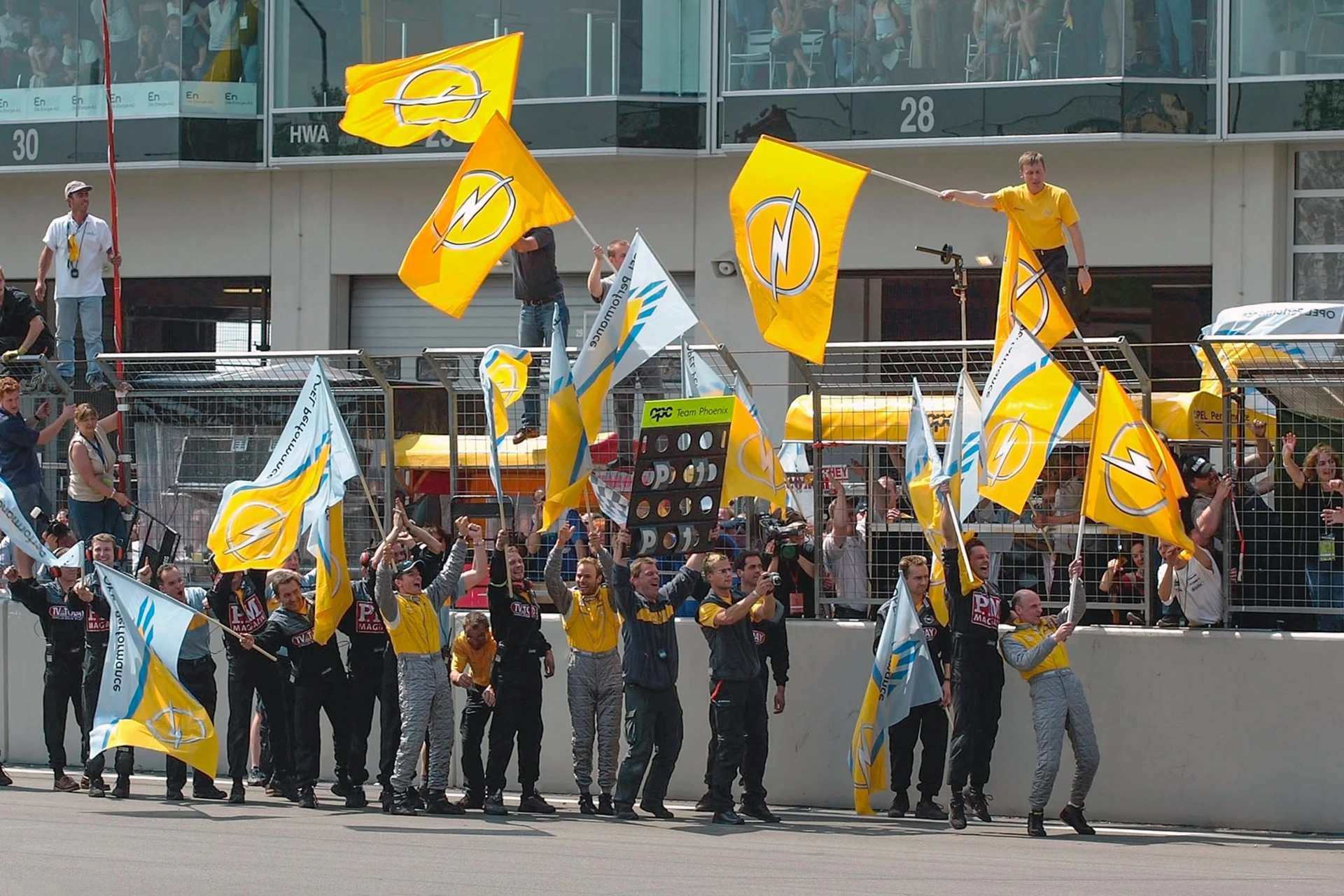 Team members and supporters in yellow and grey clothing waving large Opel flags along the pit wall at a race circuit, celebrating with raised arms.