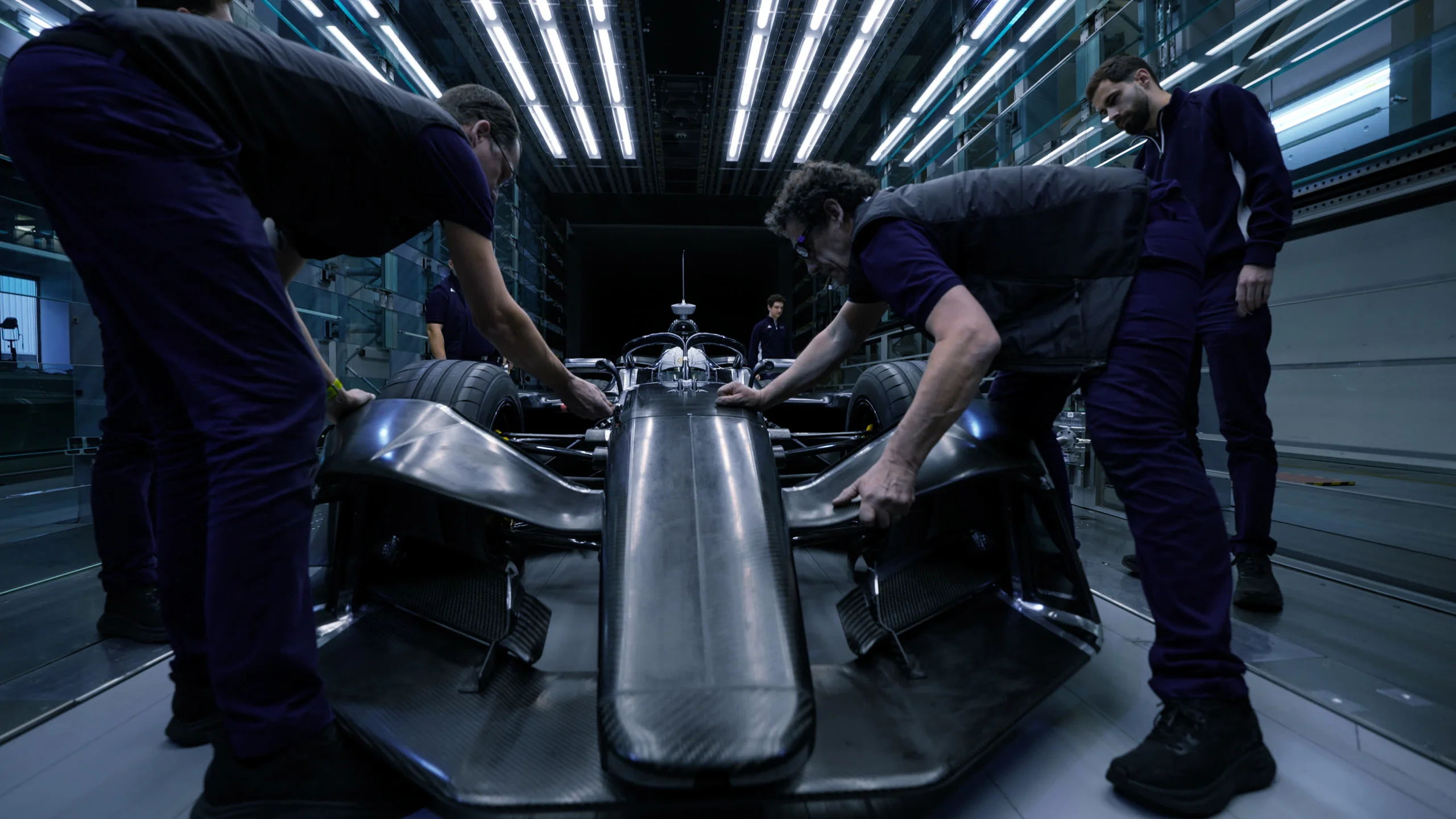Four team members in dark overalls leaning over an unpainted single-seater race car in a dimly lit garage, working on its bodywork under overhead strip lighting.