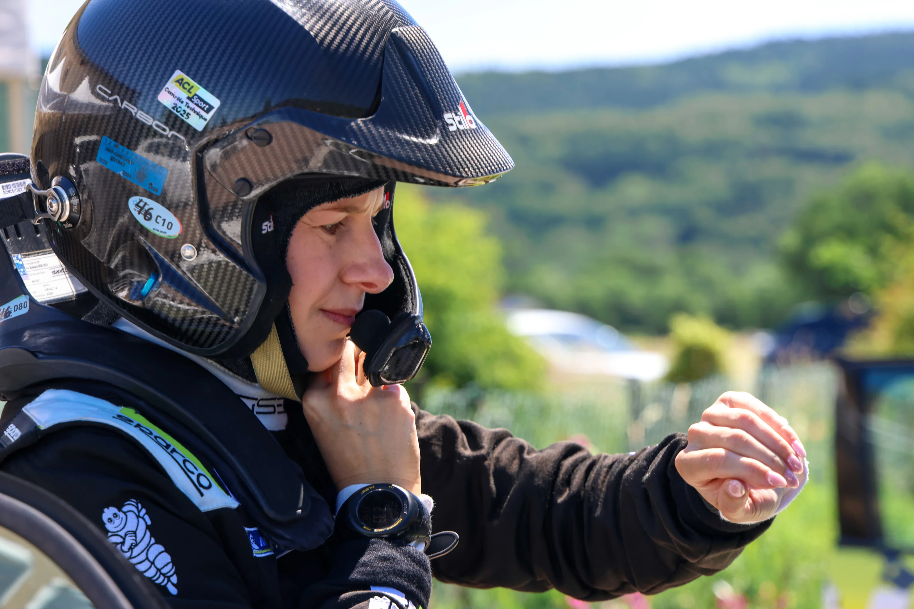 Close-up of a crew member in a black racing suit fastening the strap of a carbon fibre open-face rally helmet with a microphone boom, outdoors with green hills blurred behind.