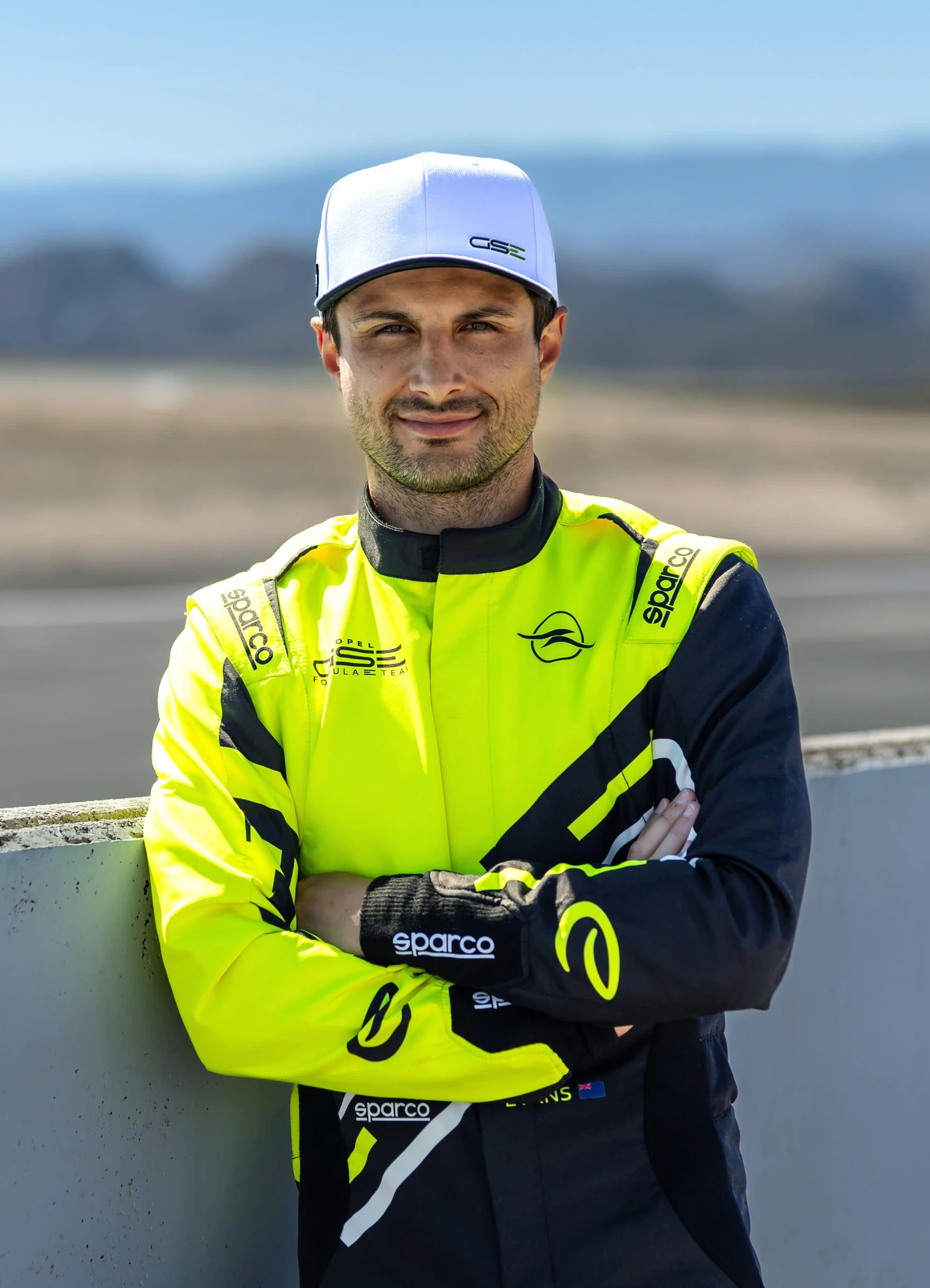 A rally driver in a neon yellow and black Opel GSE racing suit with Sparco gloves and a white GSE cap, standing with arms crossed at the edge of a race track.