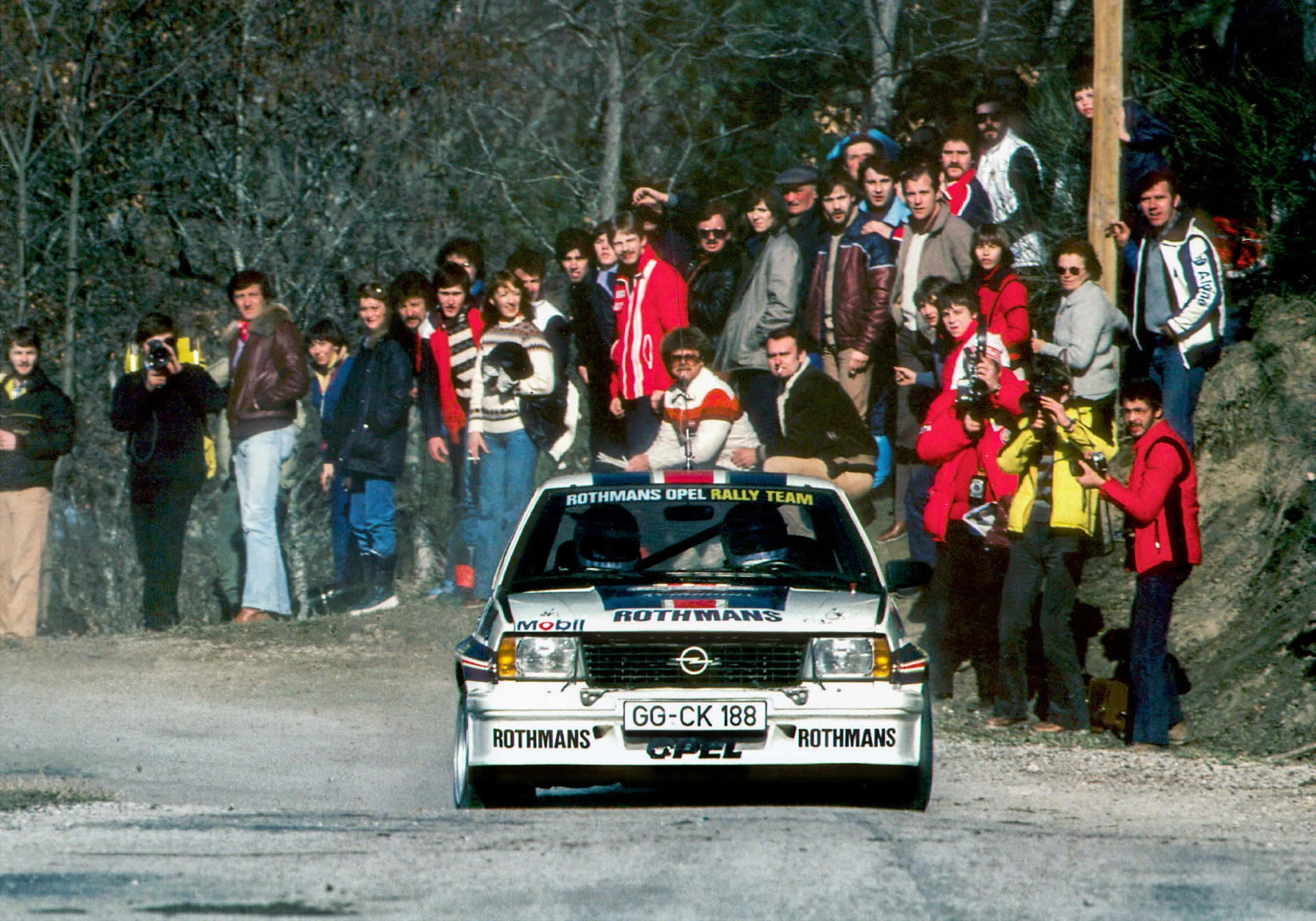 Archive photo of a white Rothmans Opel Rally Team car on a narrow gravel stage, coming towards the camera with a crowd of spectators and photographers packed on the bank behind it.