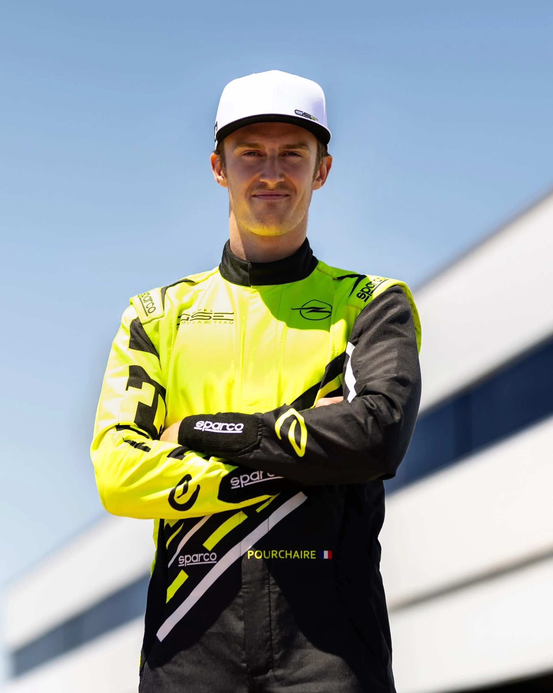 Racing driver Pourchaire in a neon yellow and black Opel GSE Formula E Team race suit and white GSE cap, standing with his arms crossed against a blue sky.