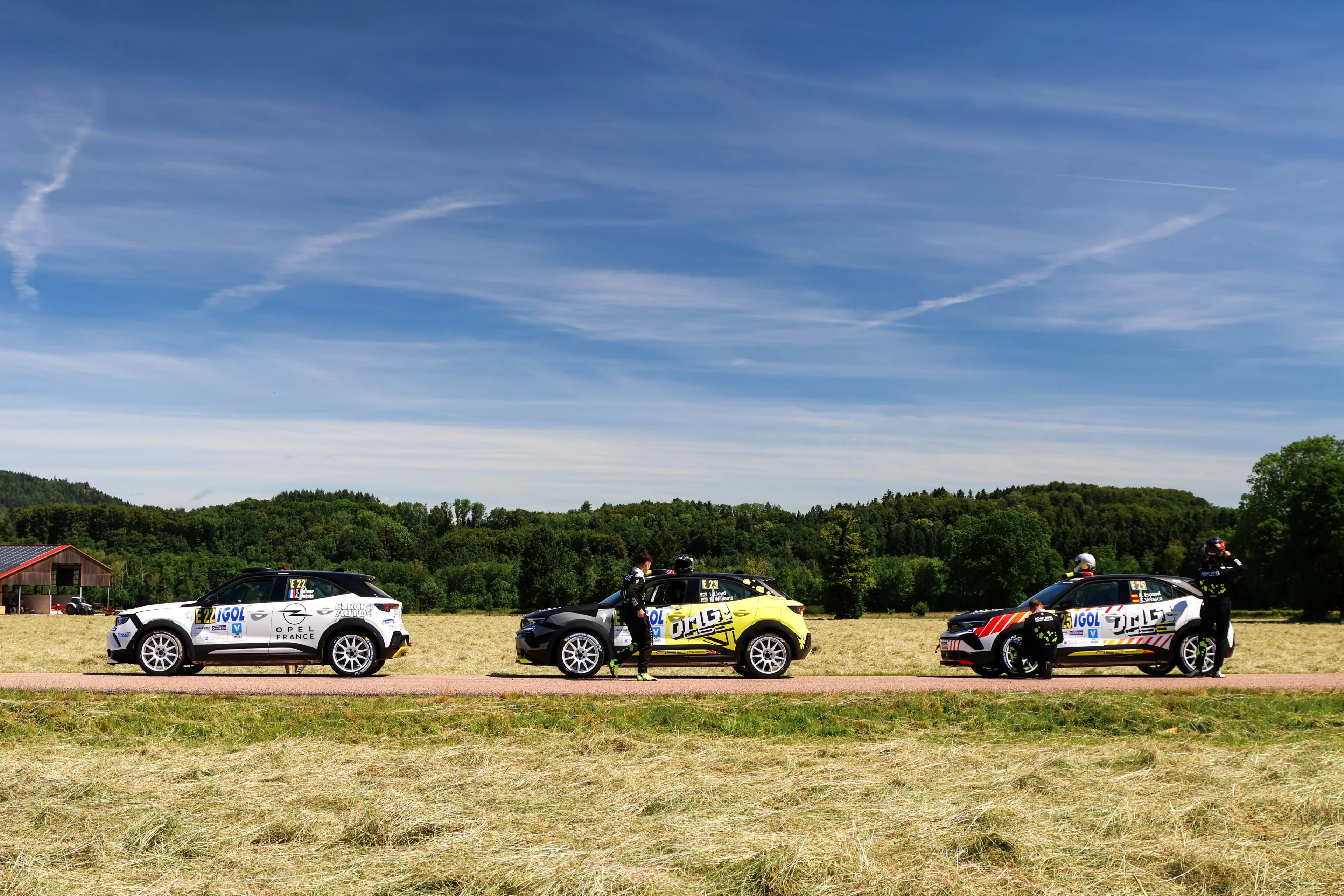 Three Opel Mokka GSE Rally cars in different liveries — white, yellow and black, and red and white — lined up along a road beside a mown field, with crew members attending to two of them and woodland behind.